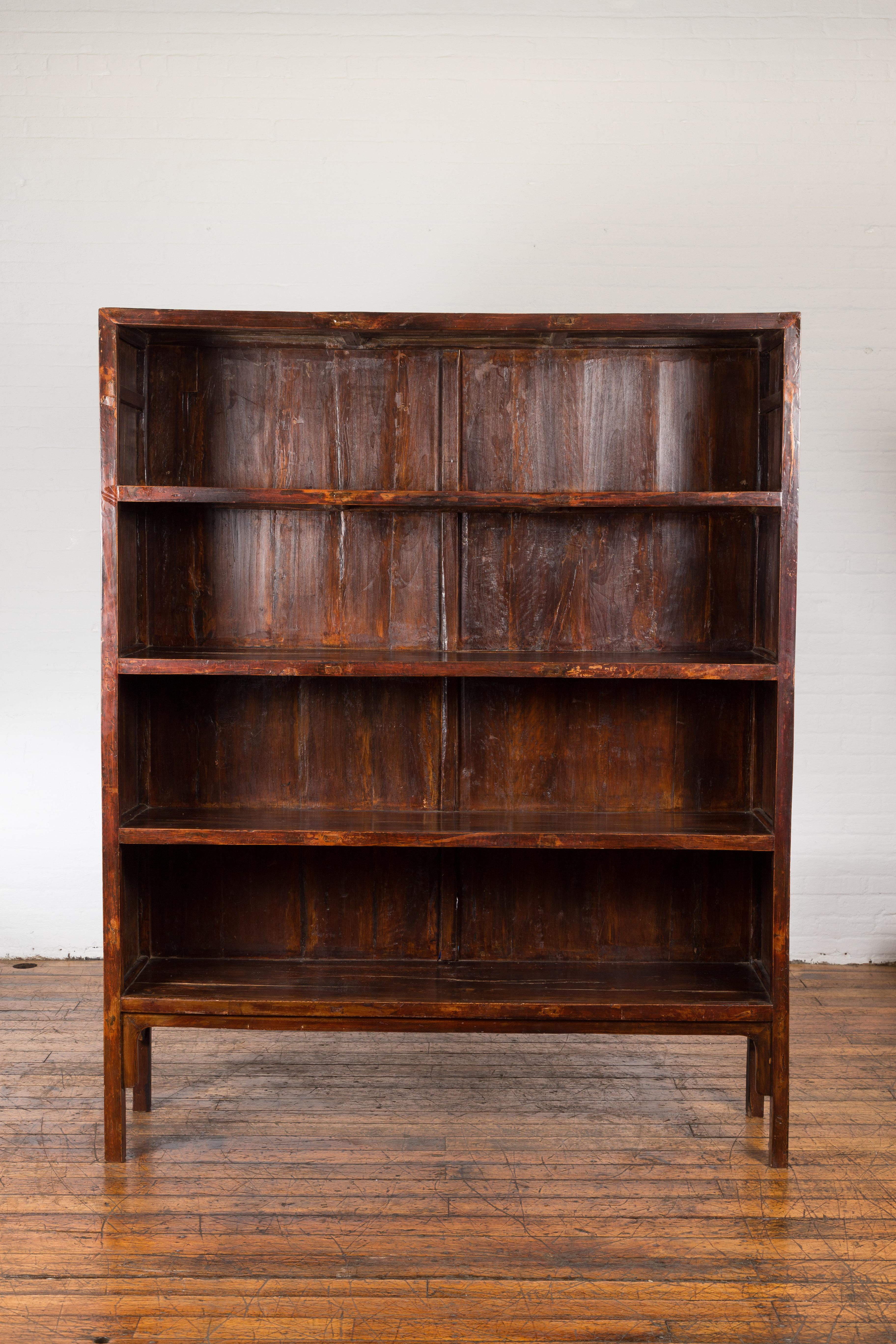 A Chinese Qing Dynasty period lacquered bookcase from the 19th century, with four shelves, straight legs, carved apron and weathered patina. Created in China during the Qing Dynasty period in the 19th century, this large bookcase features a linear