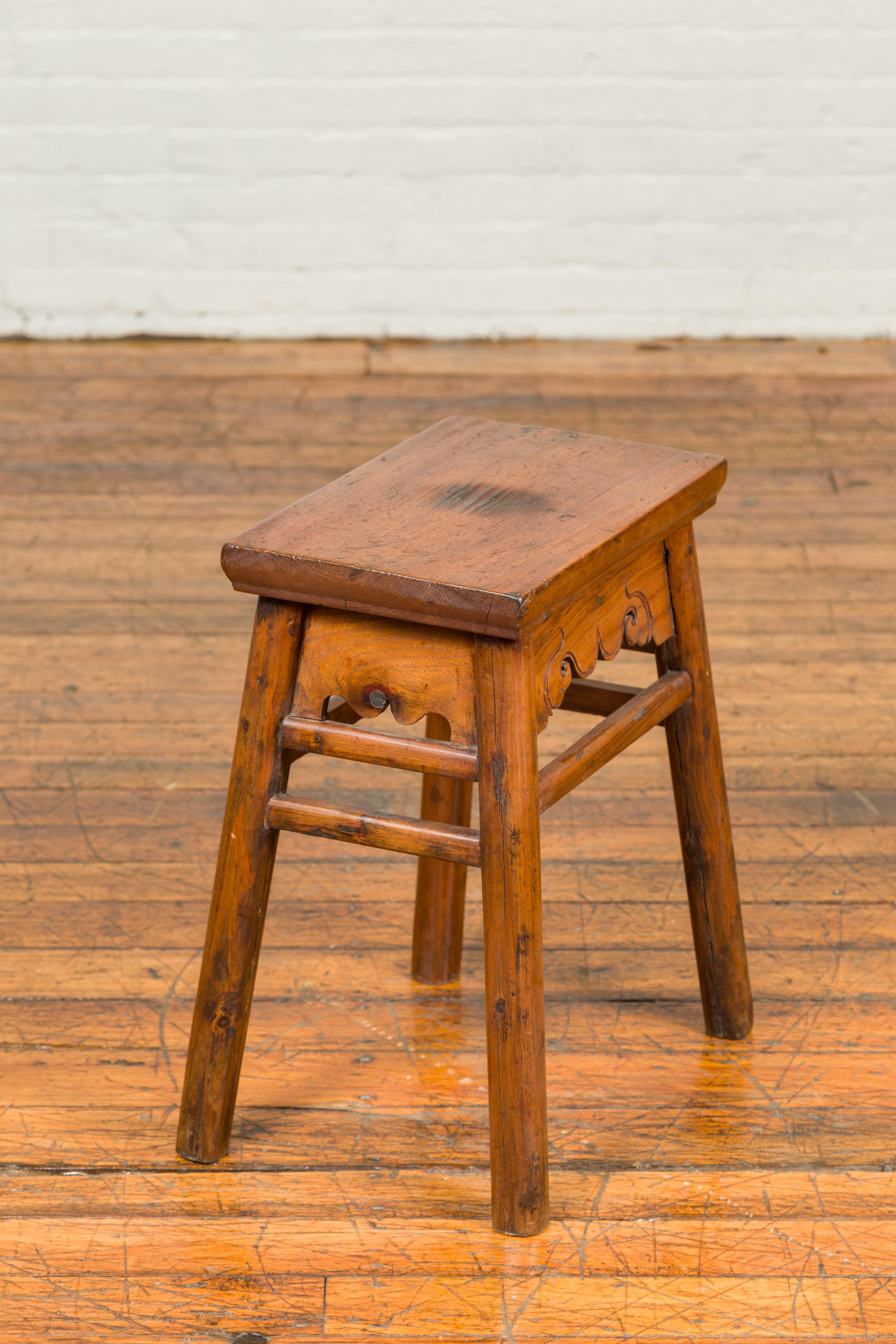 A Chinese Qing Dynasty elmwood stool from the 19th century, with carved apron and weathered top. Created in China during the Wing Dynasty, this elm stool features a rectangular top sitting above a carved apron adorned with delicate scrolls, secured