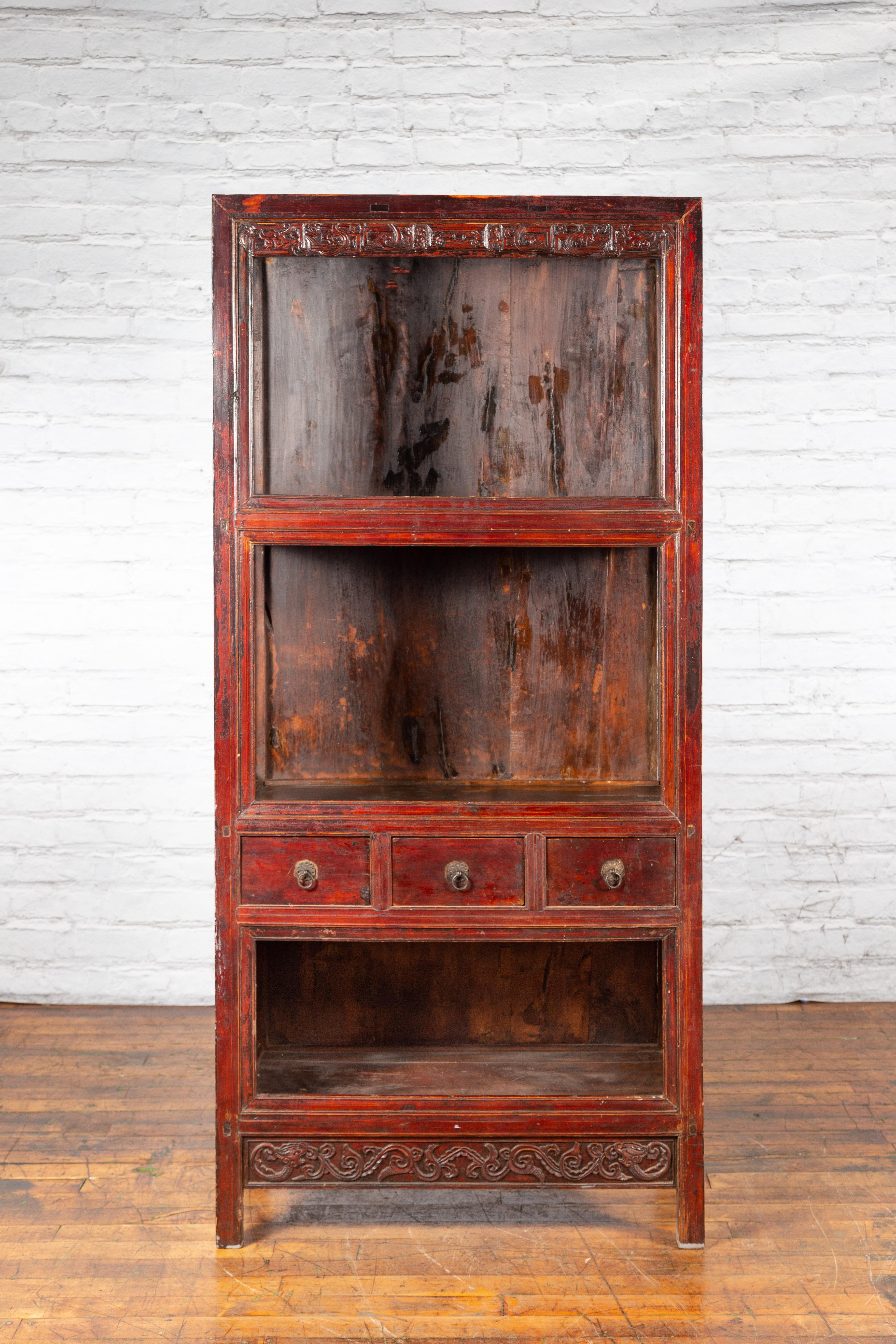A Chinese Qing Dynasty period wooden display cabinet from the 19th century, with reddish brown lacquer, open shelves, drawers and carved friezes. Created in China during the Qing Dynasty period in the 19th century, this display cabinet features a
