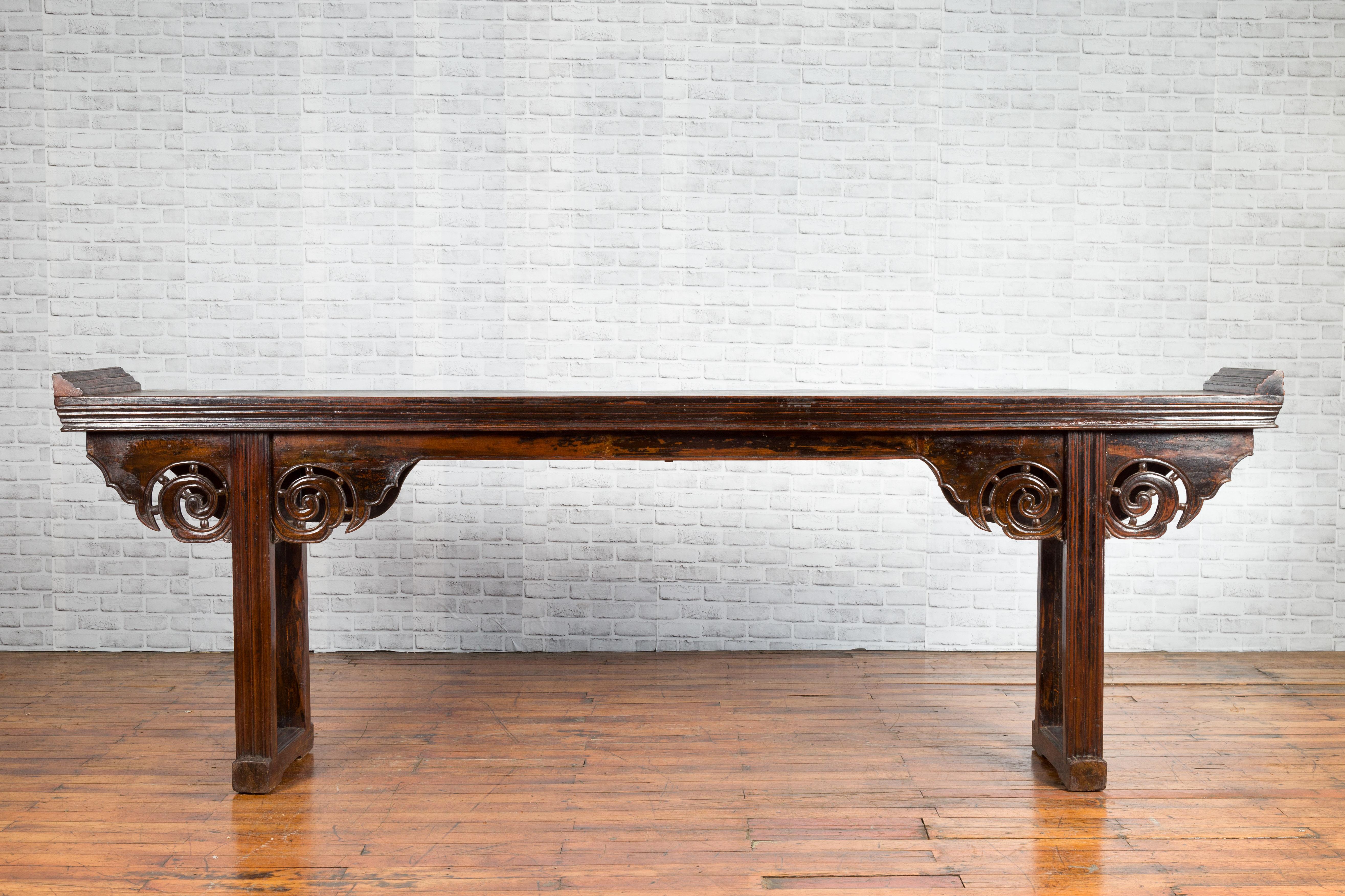 A Chinese Qing Dynasty period long altar table from the 19th century, with scrolling patterns, everted flanges and dark patina. Created in China during the Qing Dynasty, this altar console table draws our attention with its long proportions and