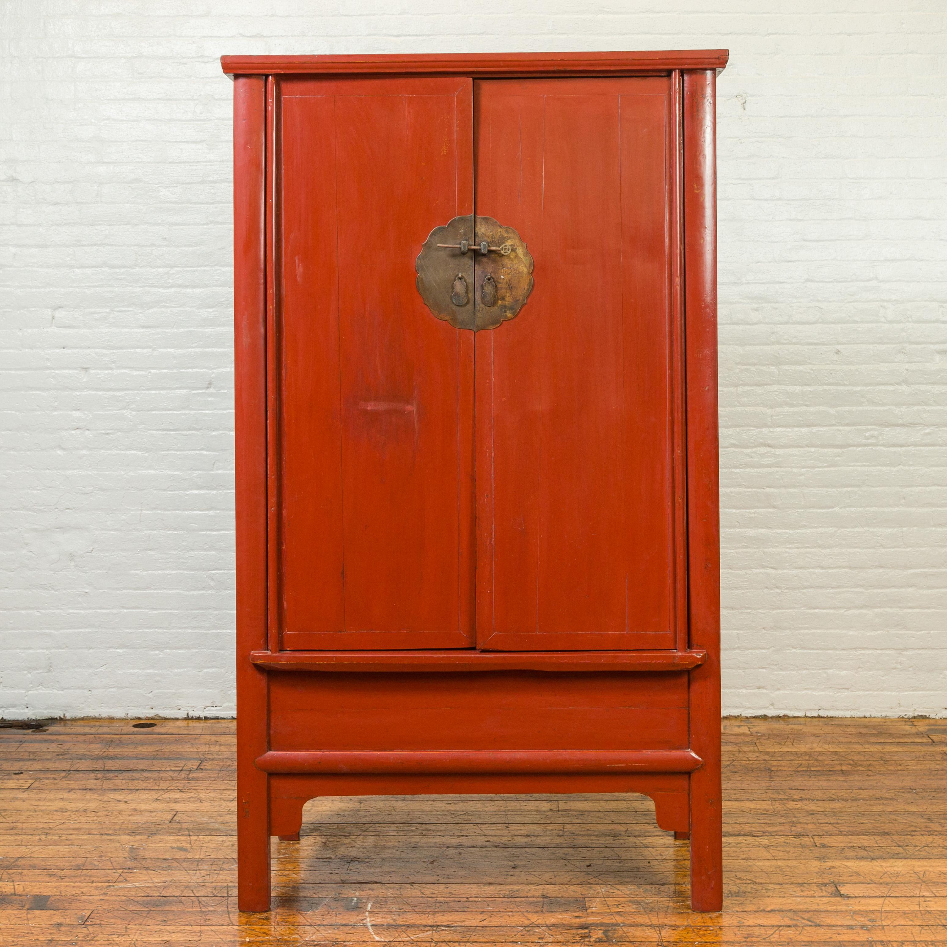 A Chinese Qing Dynasty period red lacquered armoire from the 19th century, with bronze medallion and apron. Created in China during the Qing Dynasty, this red lacquered armoire features two doors, adorned with a large bronze medallion locking thanks