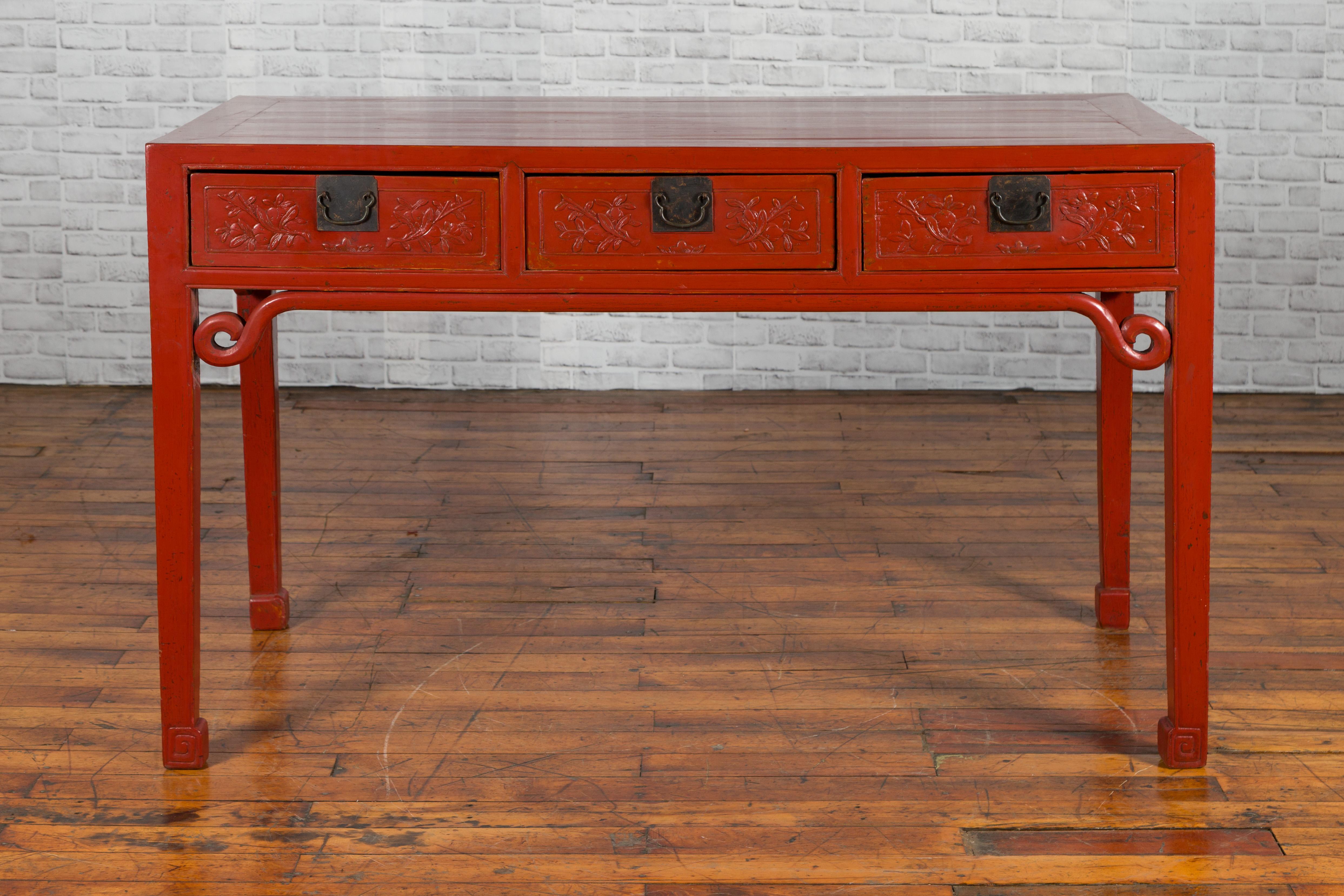A Chinese Qing Dynasty period red lacquered desk from the 19th century, with three drawers, carved stretcher and scrolling feet. Created in China during the Qing Dynasty, this red lacquered desk features a rectangular top with central board, sitting