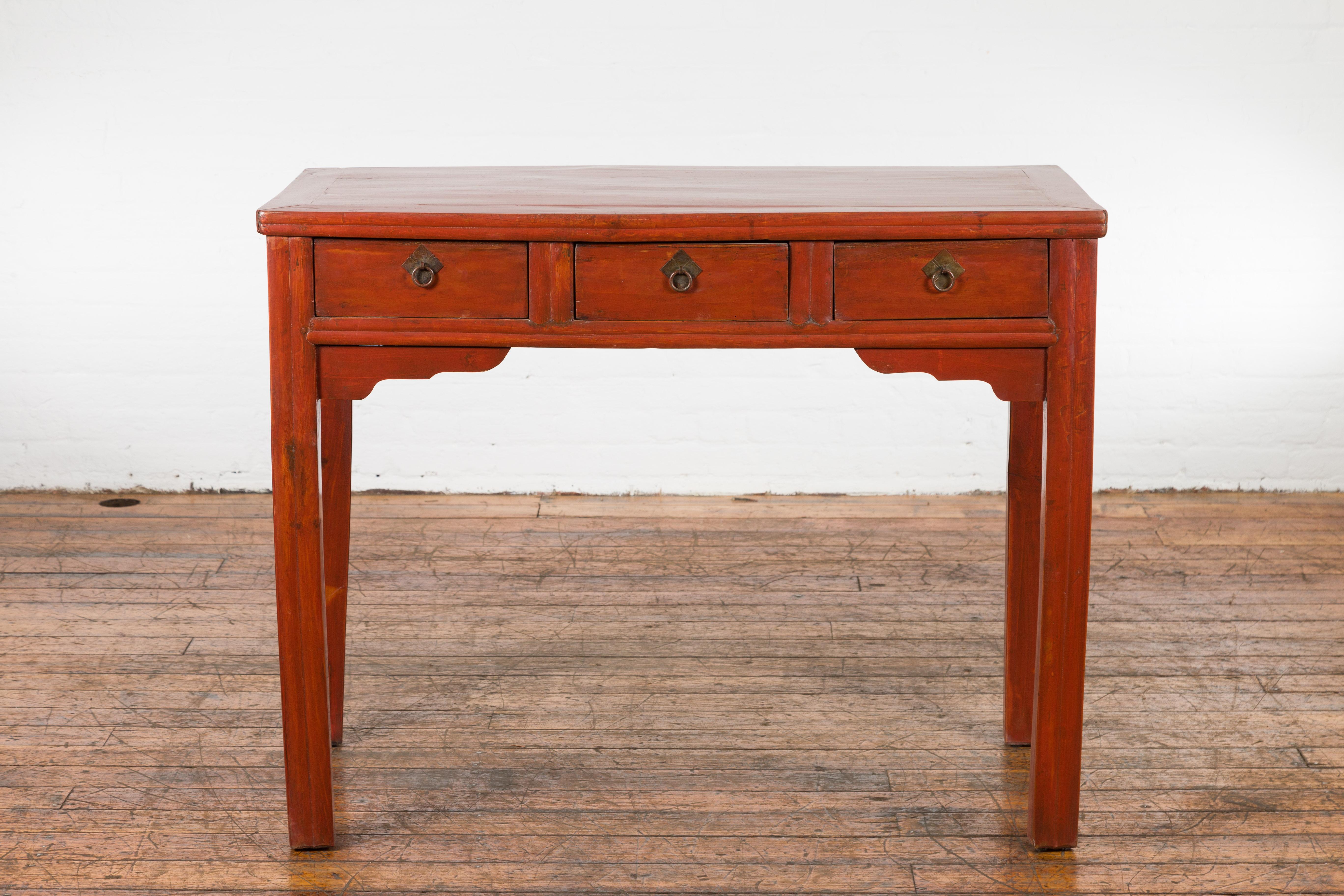 A Chinese Qing Dynasty period table from the 19th century with reddish orange lacquer, three drawers, carved spandrels and brass hardware. Created in China during the Qing Dynasty period in the 19th century, this lacquered table could be used as a