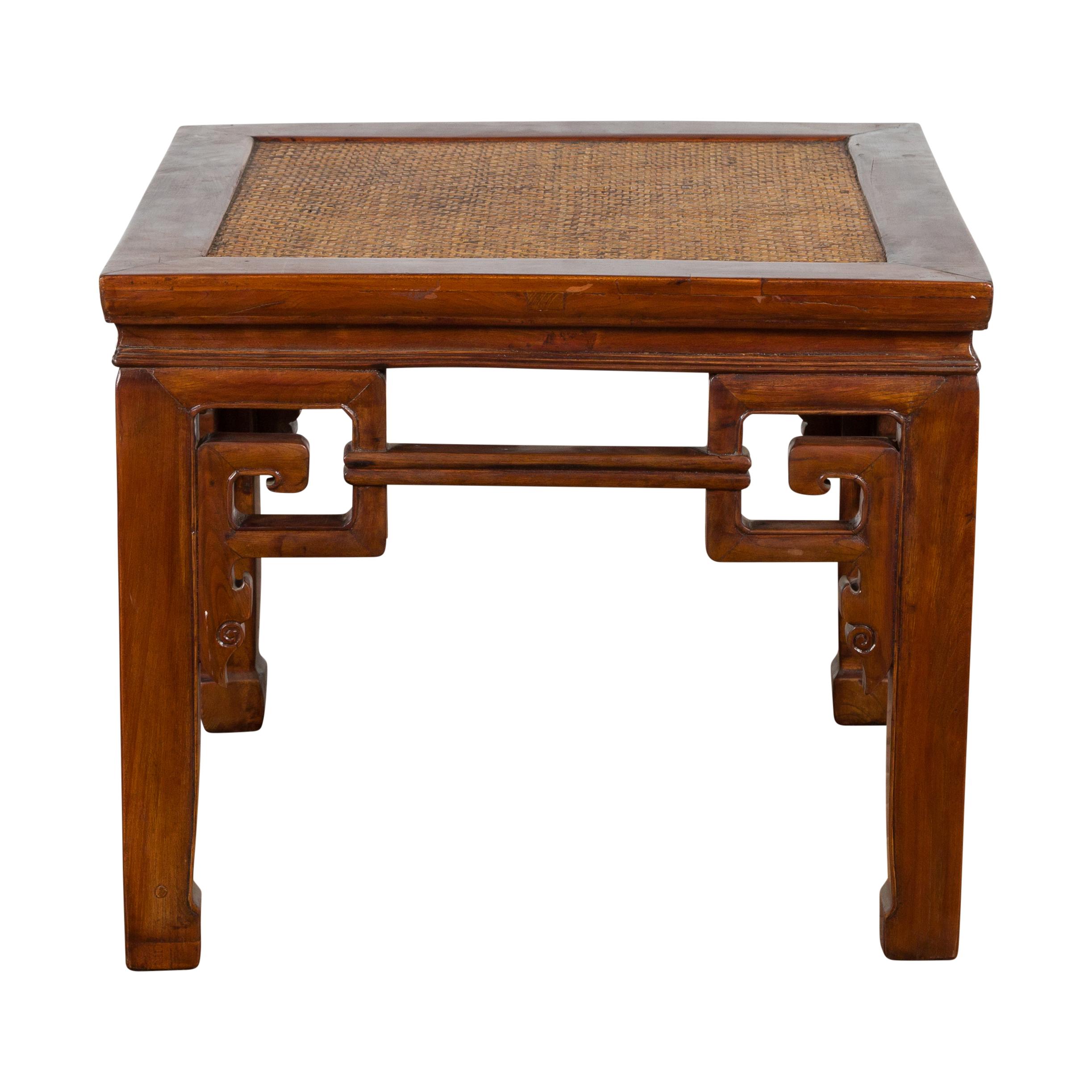 Petit tabouret ancien en bois d'orme chinois d'époque dynastie Qing du 19ème siècle avec écoinçons à volutes sculptées, plateau en rotin tressé et pieds en sabot de cheval, pouvant également servir de table à boissons. Créé en Chine pendant la