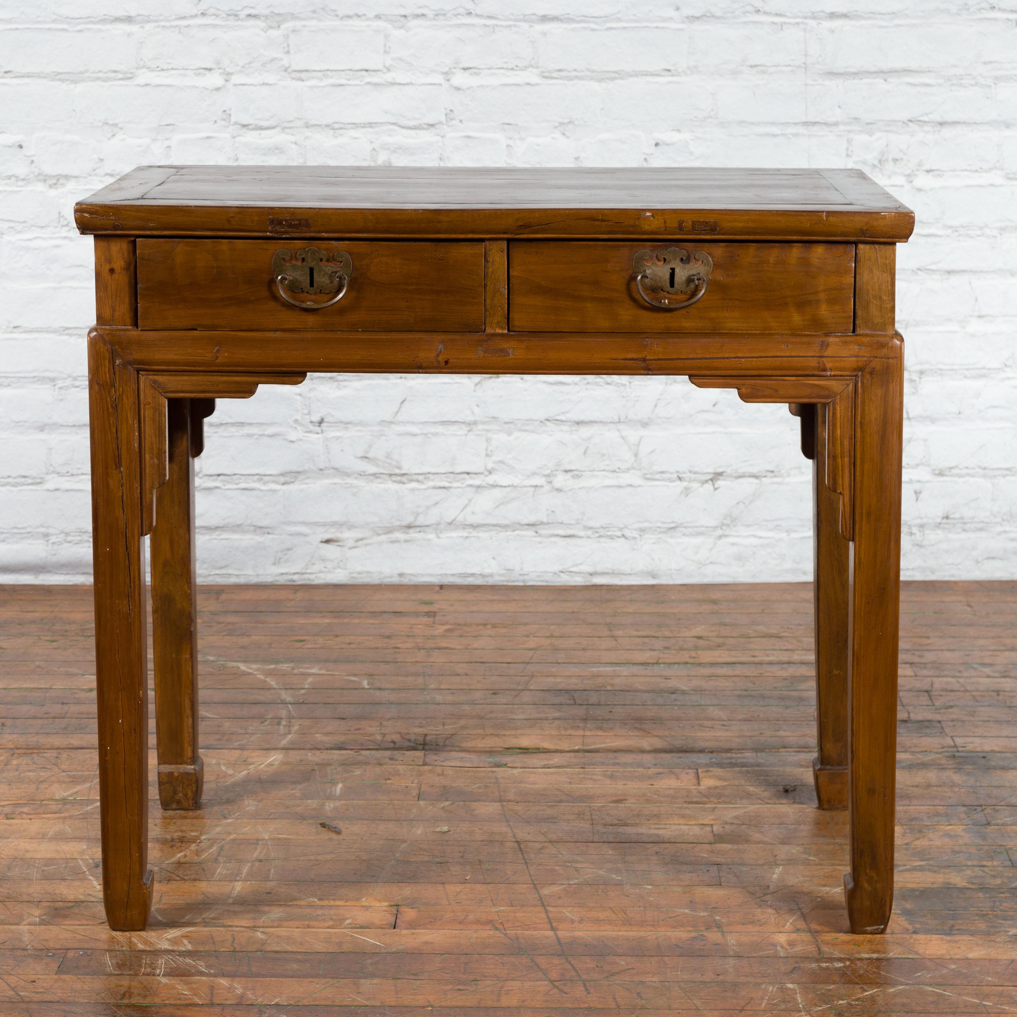 A Chinese Qing Dynasty period wooden desk from the 19th century with two drawers, horse hoof legs, carved spandrels and butterfly bronze hardware. Created in China during the Qing Dynasty period in the 19th century, this wooden desk features a