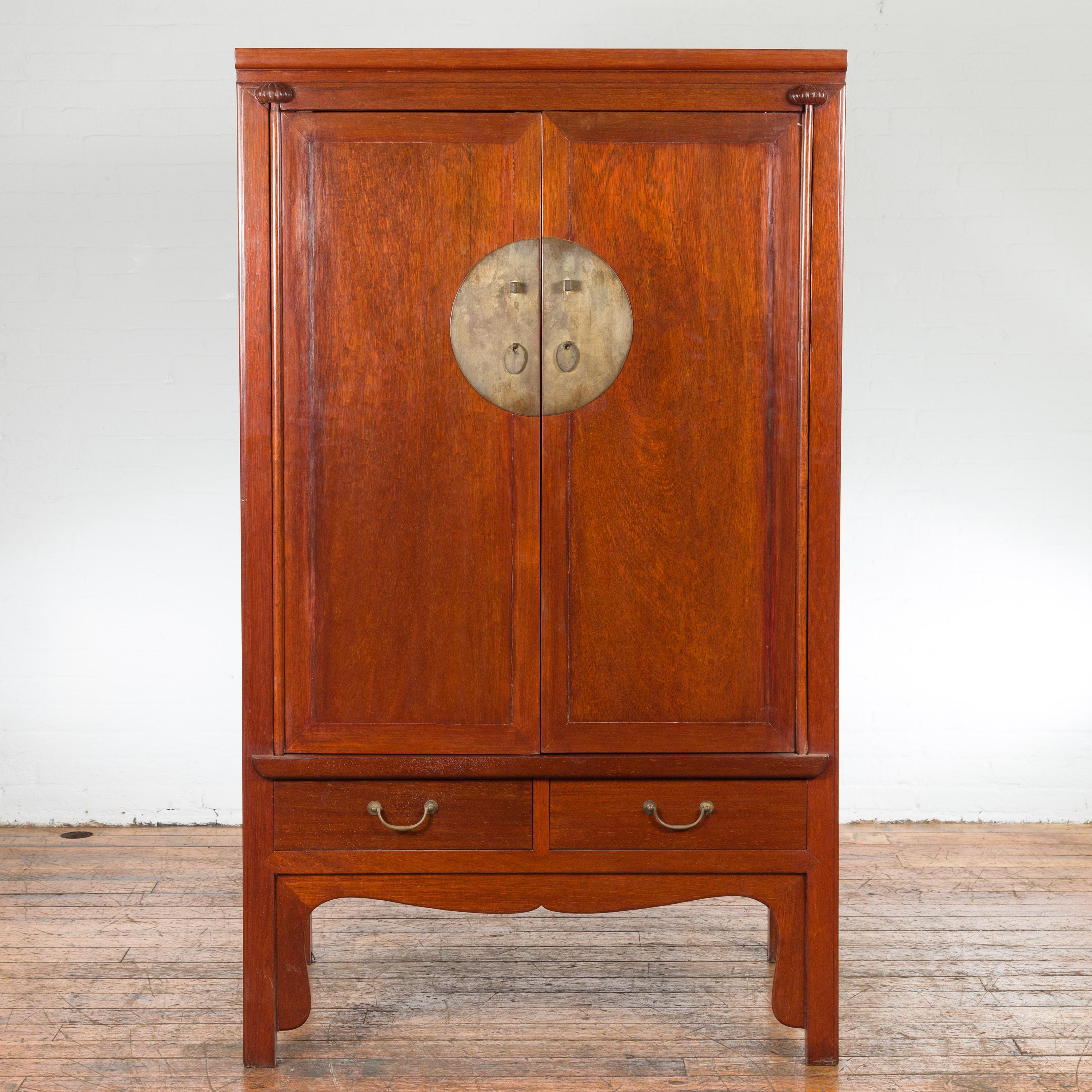 A Chinese Qing Dynasty period brown armoire from the 19th century, with brass round medallion, reconfigured pocket doors, two drawers and carved apron. Created in China during the Qing Dynasty period in the 19th century, this armoire features a