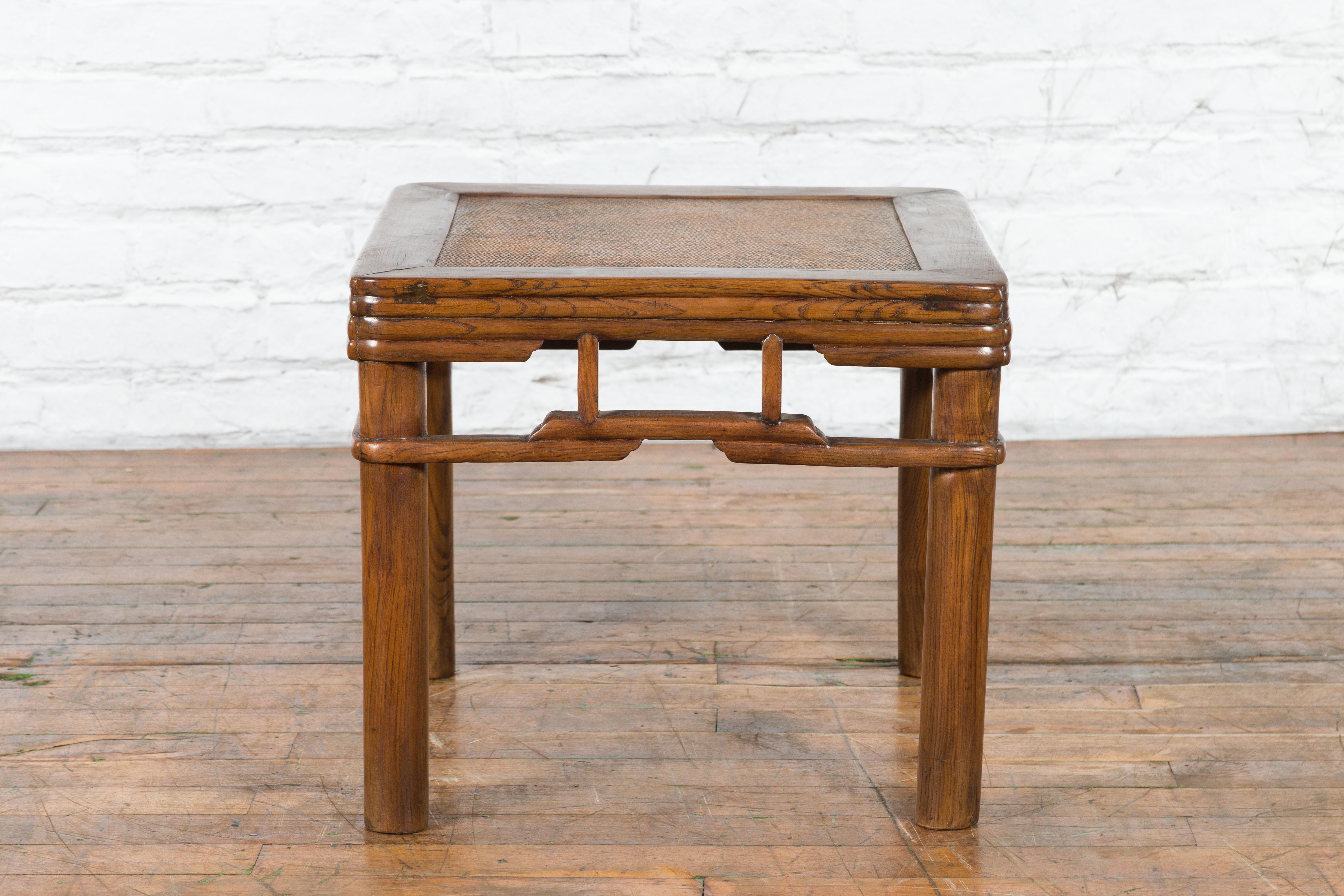 A Chinese Qing Dynasty period elm wood side table from the early 20th century with woven rattan top inset, reeded apron, and pillar strut motifs. Created in China during the Qing Dynasty period in the early years of the 20th century, this elm wood