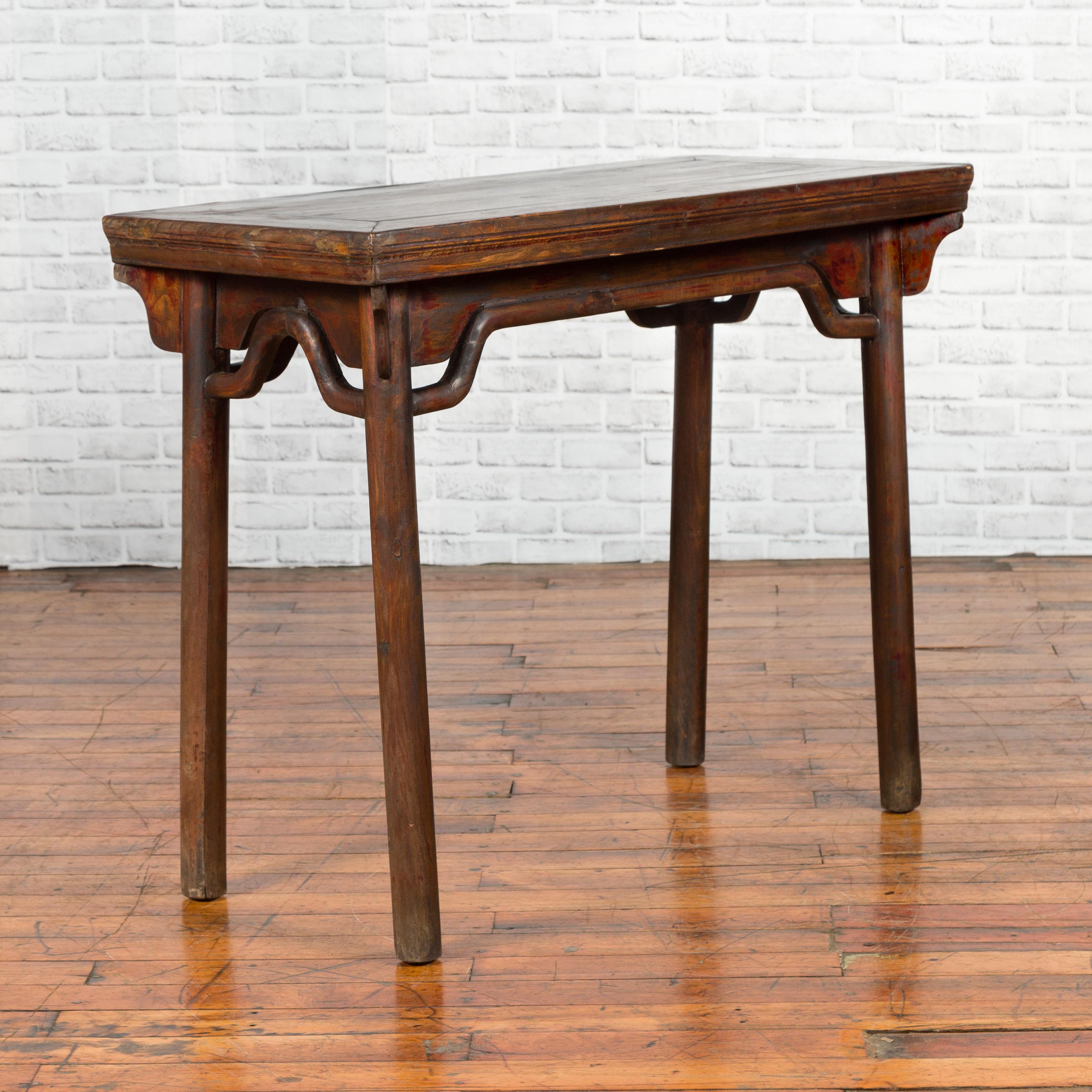 A Chinese Qing Dynasty elmwood wine table from the 19th century, with humpbacked apron and carved spandrels. Created in China during the 19th century, this elm wine table features a rectangular top with central board, sitting above a humpbacked