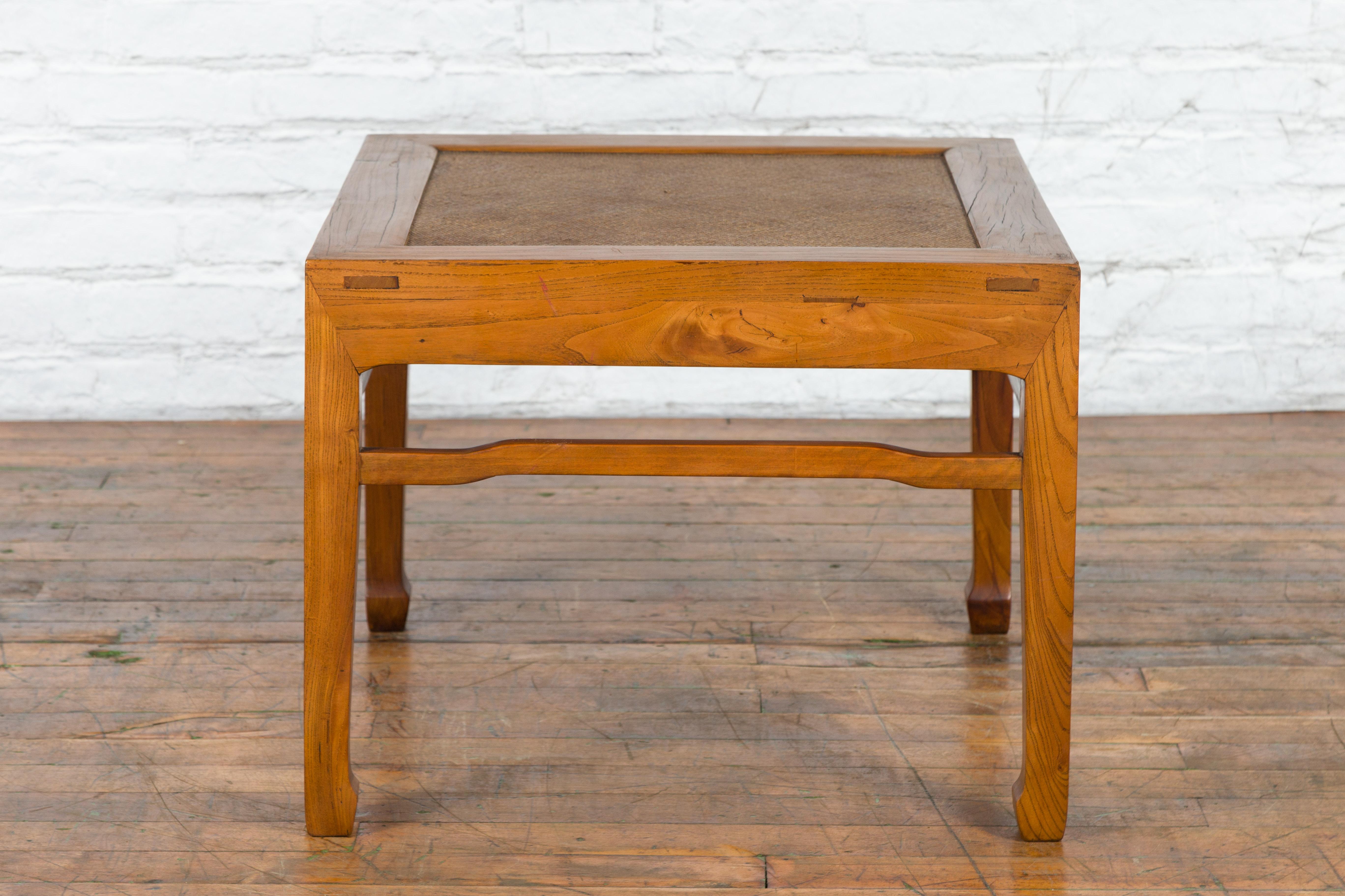 A Chinese Qing Dynasty period elm wood side table from the 19th century, with woven rattan top, humpback stretchers and horse hoof feet. Created in China during the Qing Dynasty period in the 19th century, this elm wood side table features a square