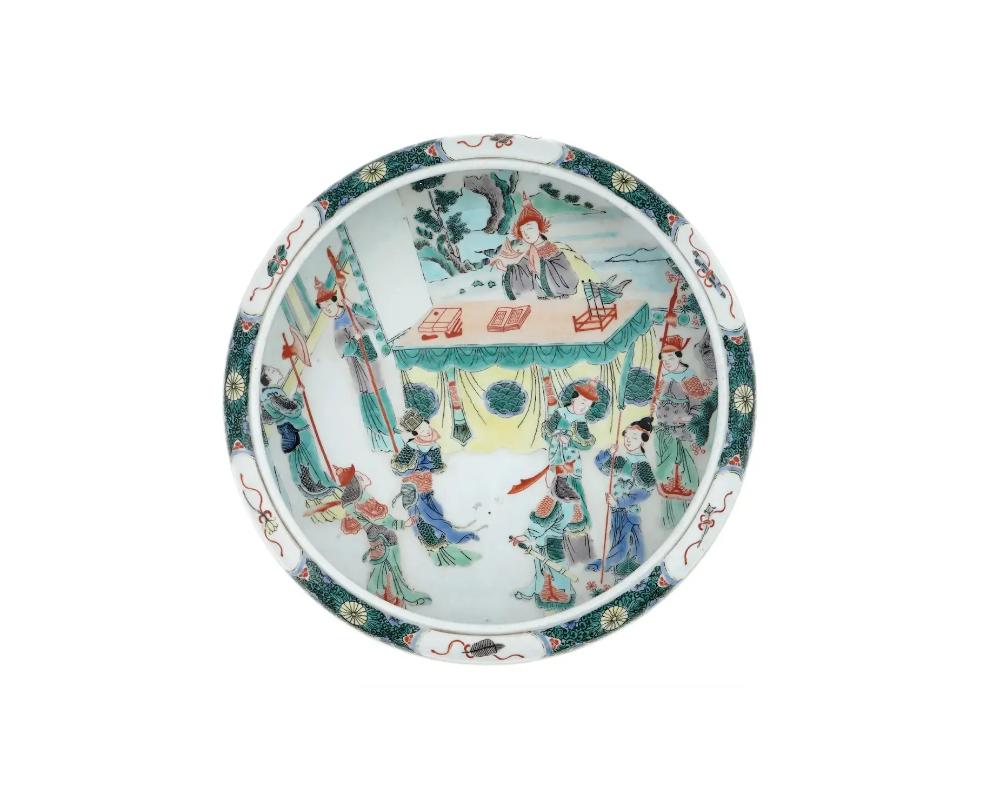 An antique Chinese glazed white porcelain dish. Late Qing dynasty, before 1912. Round deep plate with high rims. The piece is decorated with hand-painted polychrome Famille rose decor. The center of the plate depicts a multi-figure scene with