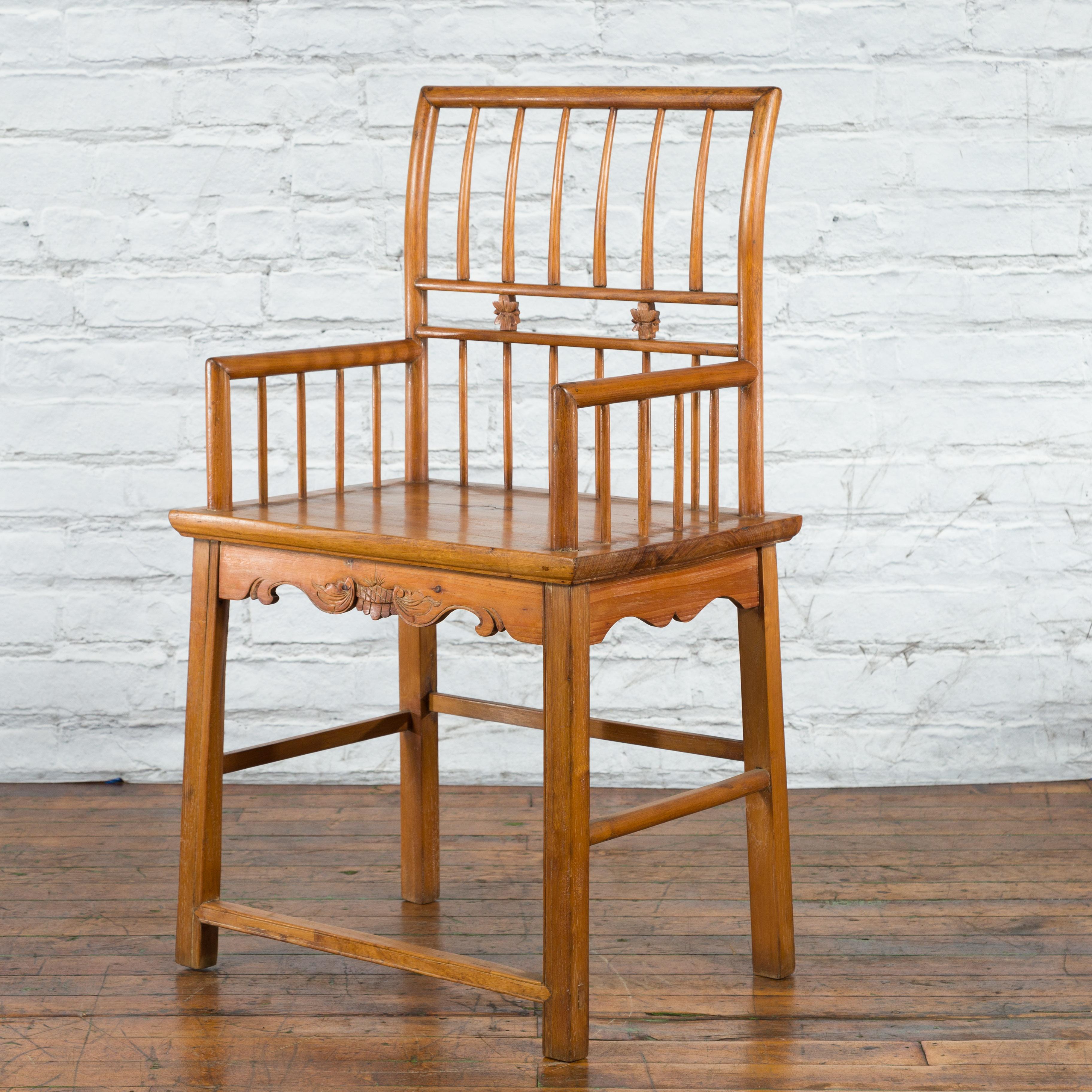 A Chinese Qing Dynasty period elm wood armchair from the 19th century with hand-carved apron and floral motifs. Created in China during the Qing Dynasty in the 19th century, this elm wood armchair features a slightly out-scrolling stick back,