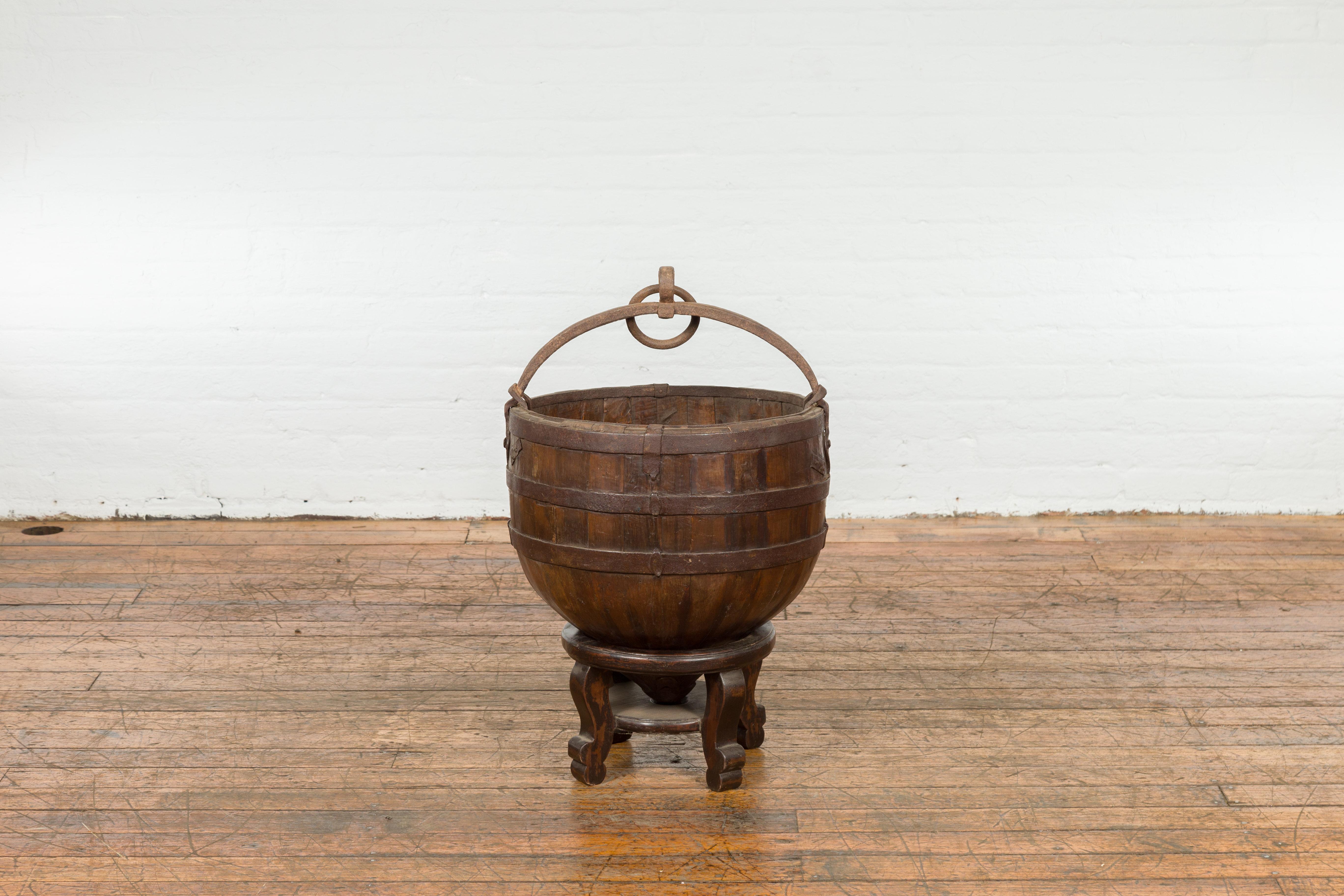 A rustic Chinese Qing Dynasty period wooden rice bucket from the 19th century, with circular shape, iron handle, custom carved stand and weathered appearance. Created in China during the Qing Dynasty period in the 19th century, this rice bucket has