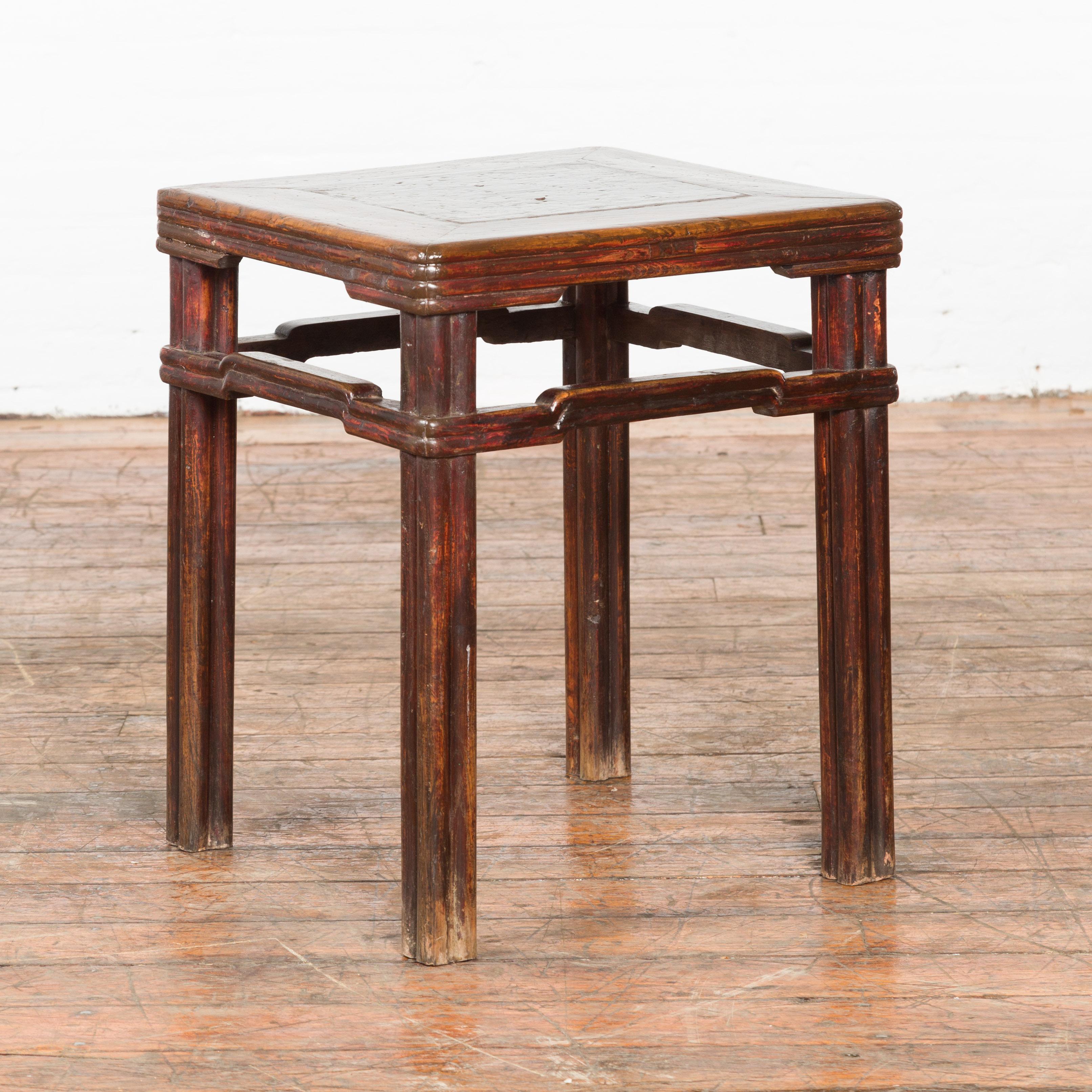 A Chinese Qing Dynasty period side table from the 19th century, with humpback stretchers and red undertone. Created in China during the Qing Dynasty, this side table features a square top with central board, sitting above a reeded apron. Raised on