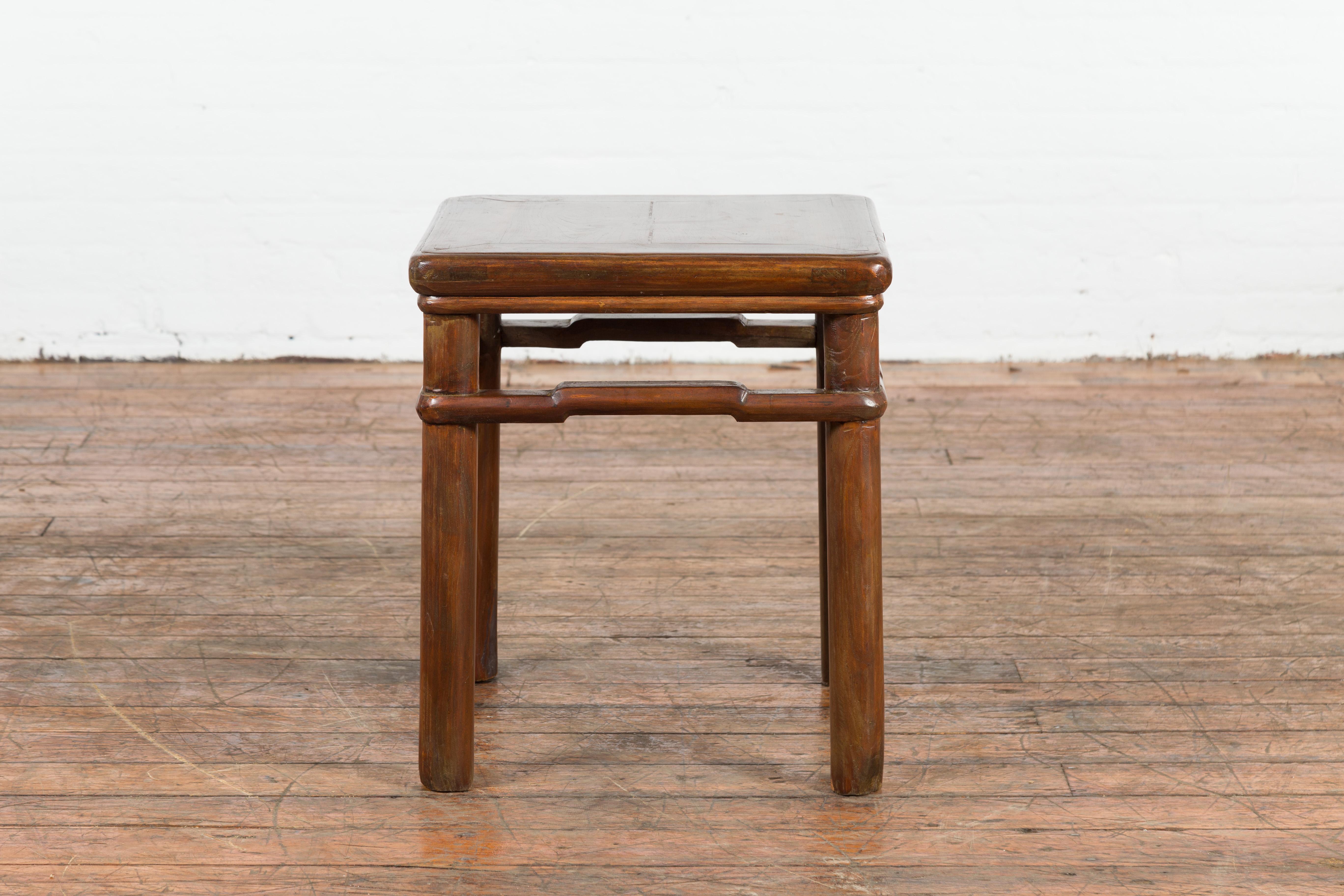 A Chinese Qing Dynasty period antique side table from the 19th century, with humpback stretchers. Created in China during the Qing Dynasty, this side table features a square top with central board, raised on four cylindrical legs connected to one