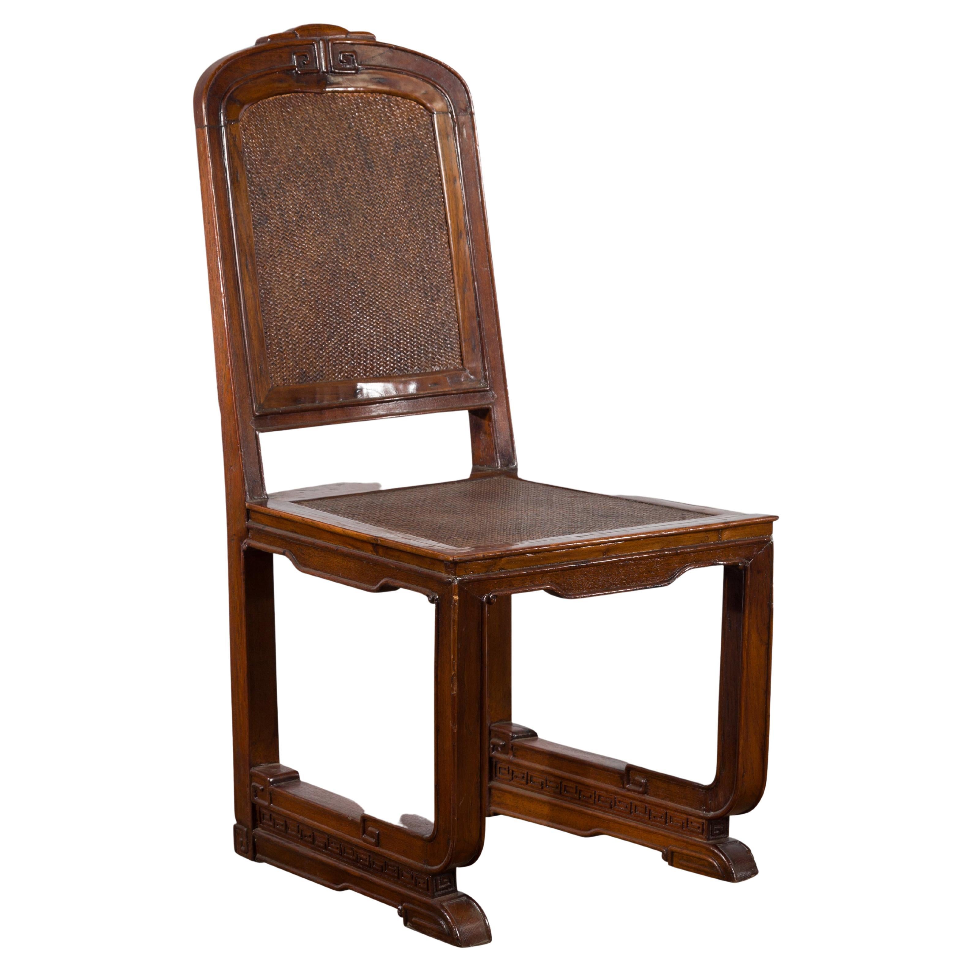 A Chinese Qing Dynasty period dark brown lacquer wooden side chair from the 19th century with rattan seat and back, carved scrolls and meander motifs. Created in China during the Qing Dynasty period in the 19th century, this wooden side chair
