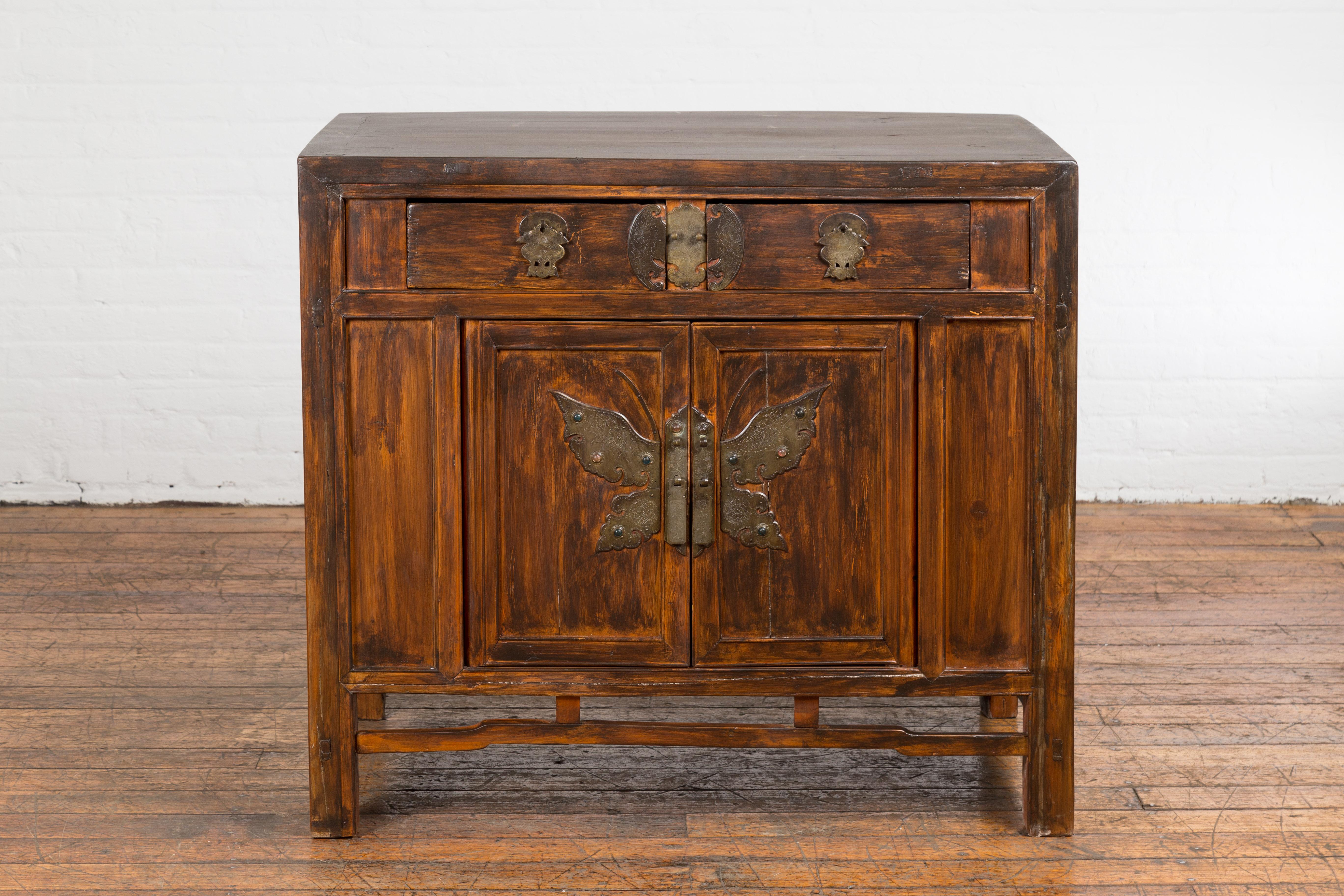 A Chinese Qing Dynasty cabinet from the 19th century with brass butterfly hardware, two drawers over a pair of double doors and distressed brown finish. Imbued with the rich heritage of Chinese craftsmanship, this 19th century Qing Dynasty cabinet