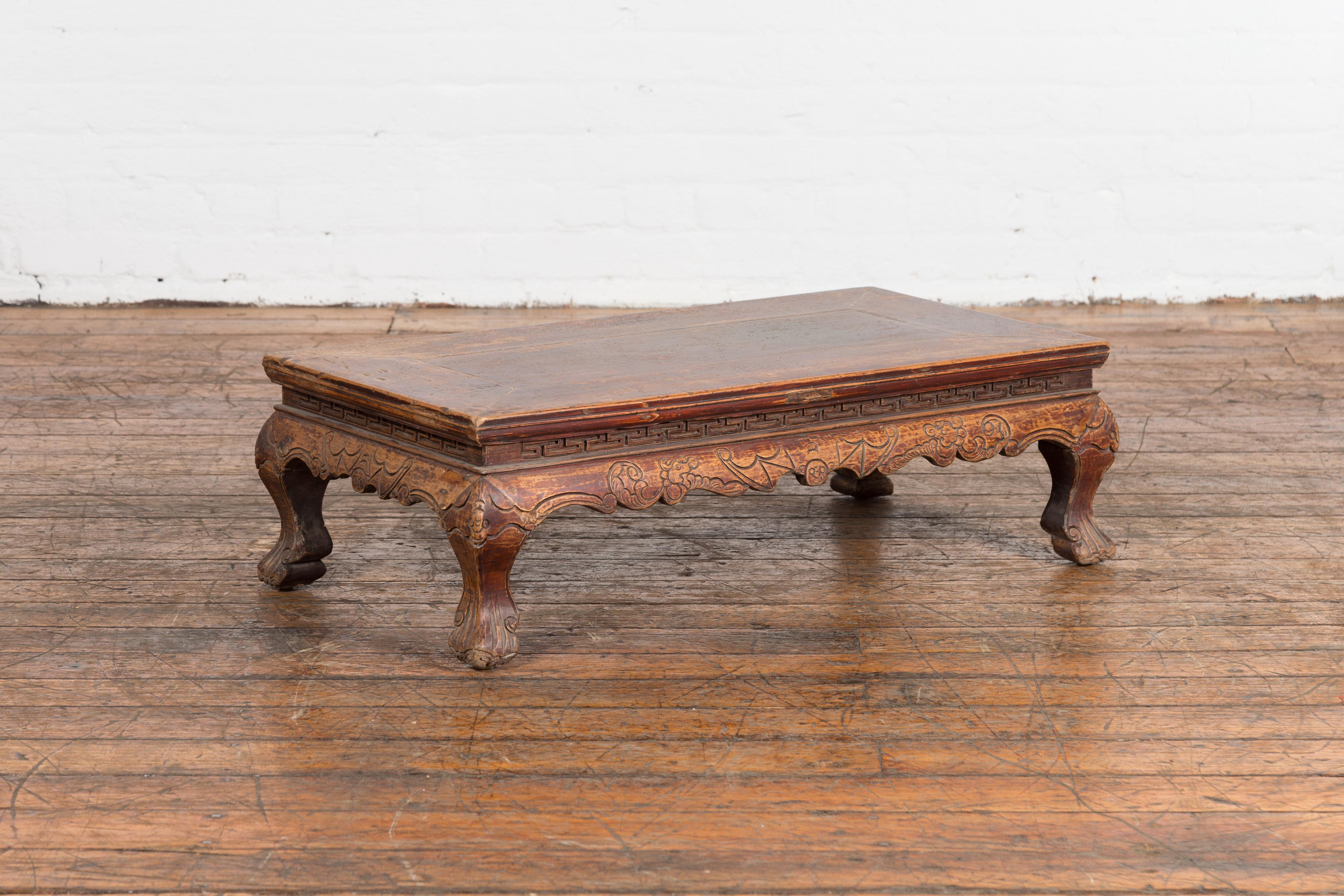 A Chinese Qing Dynasty period low Kang coffee table from the 19th century with carved meander motifs, bats, scrolling clouds and cabriole legs. Dating back to the 19th century, this Chinese low Kang coffee table is a stunning relic from the Qing
