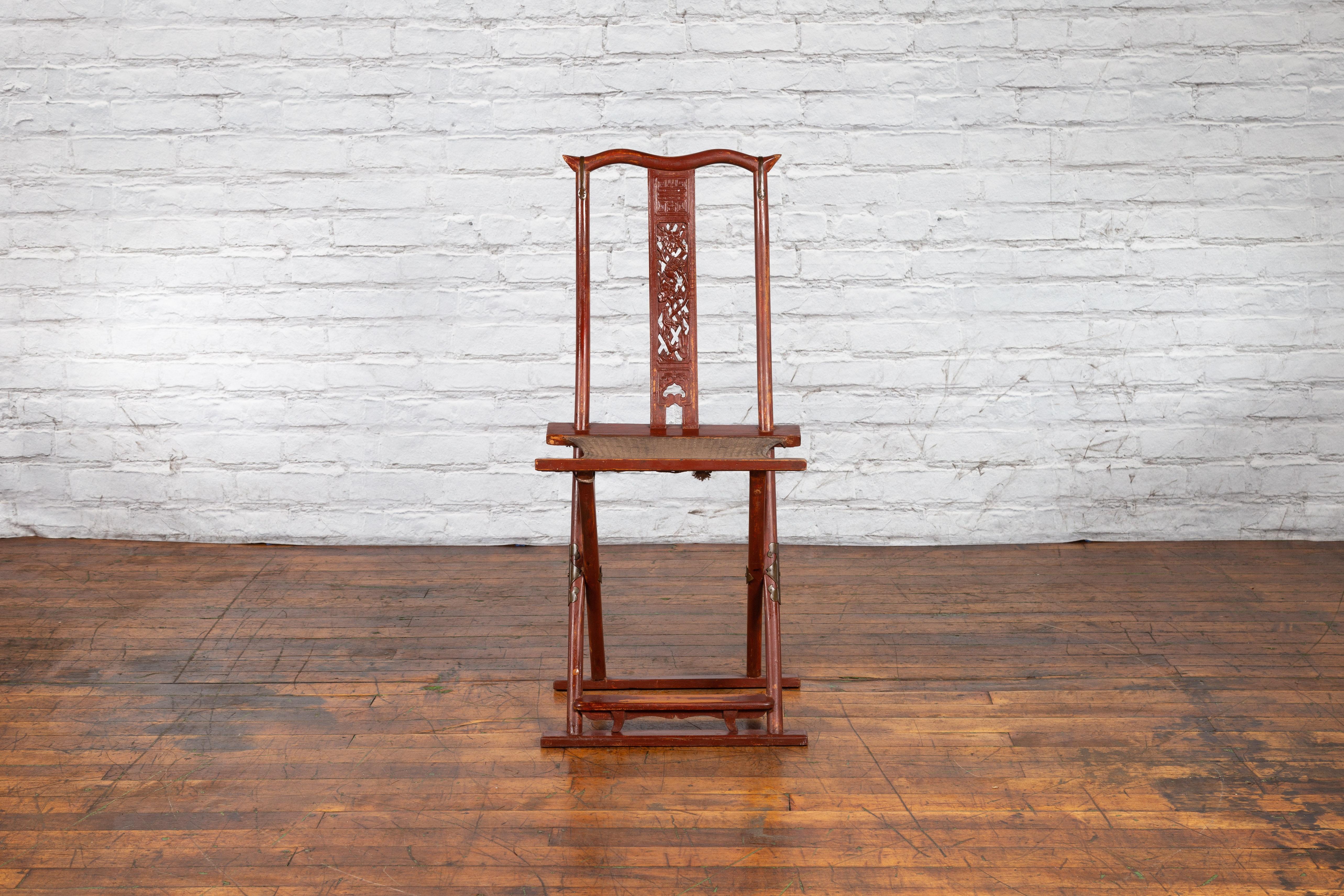 An antique Chinese Qing Dynasty period red lacquered folding traveller’s chair from the 19th century, with carved birds and foliage, scalloped footrest and woven fabric. Created in China during the Qing Dynasty period in the 19th century, this