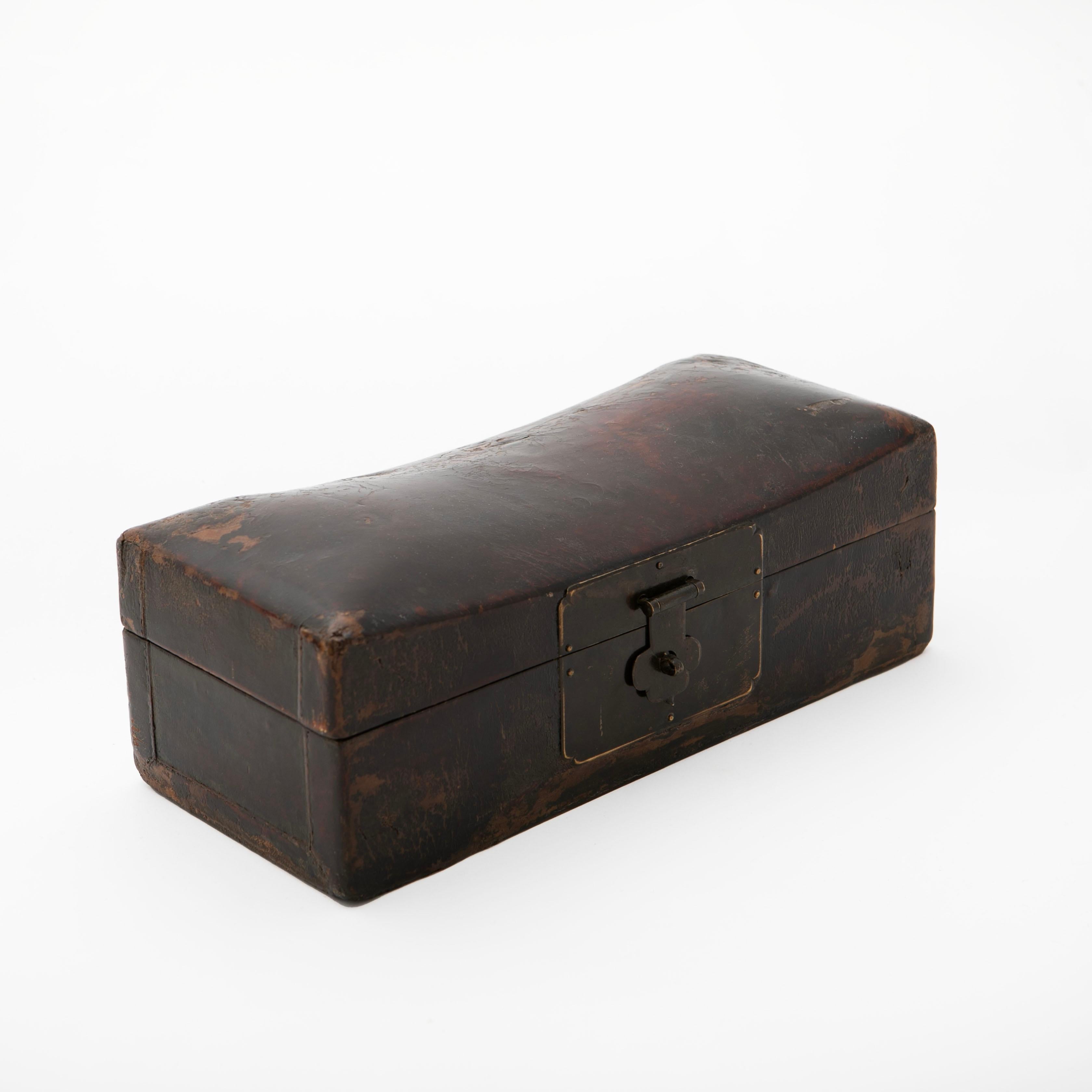 An antique Qing dynasty wooden 'pillow box' covered with leather and a burgundy lacquer finish.
Features a curved top and a patinated brass lock plate.

Originally used as a headrest to store valuables and important documents while sleeping. 

China