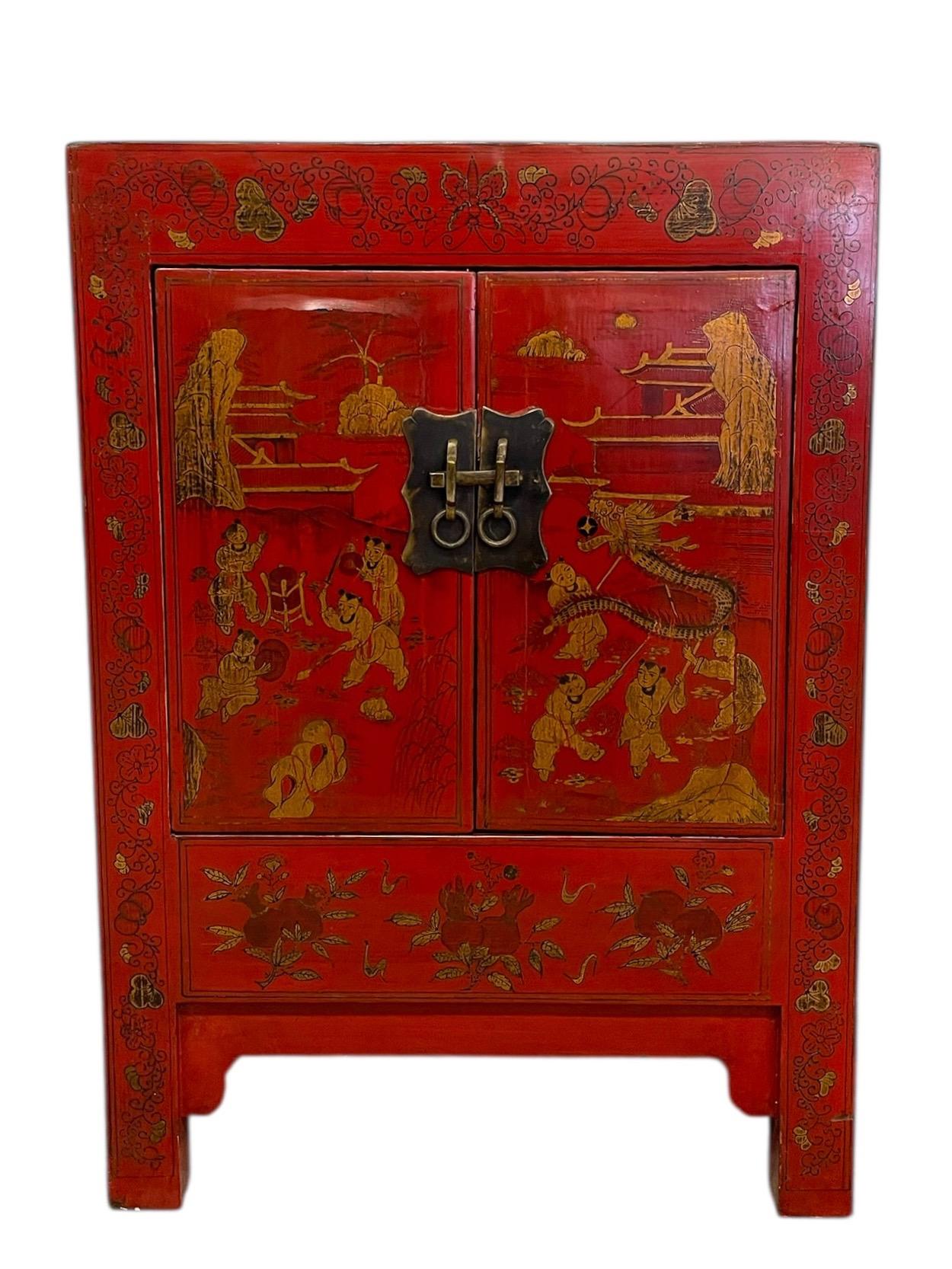 This Chinese red cabinet features finely detailed gilt decoration depicting festive scenes, pagoda architecture, and a traditional dragon dance motif. The front panels are richly adorned with figures in landscape settings, surrounded by a decorative