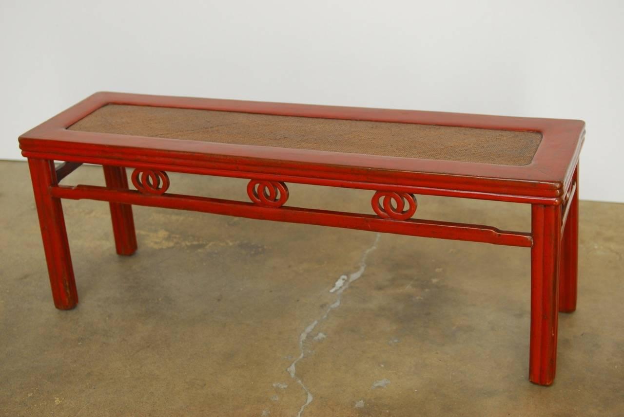 Stylish Chinese red lacquered carved bench featuring a raffia covered seat. Beautifully carved with reeded legs and sides. The aprons have a double circle motif attached to humpback stretchers on each side. The solid wood is finished in a China red