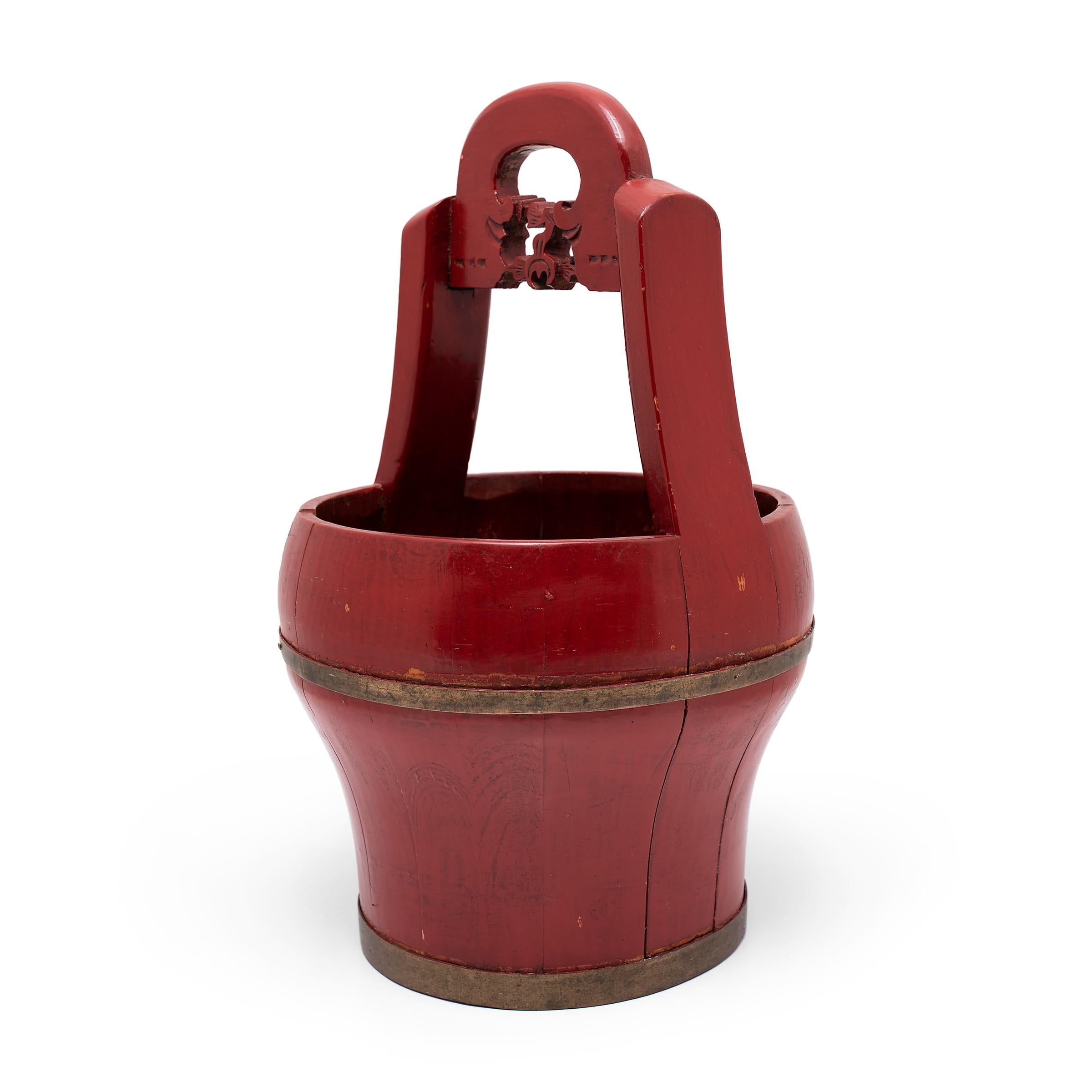 This red lacquer bucket dates to the late 19th century and was originally used as a portable container for carrying food, water, and other goods for long distances. The tall carved handle allowed the container to be placed on a carrying pole