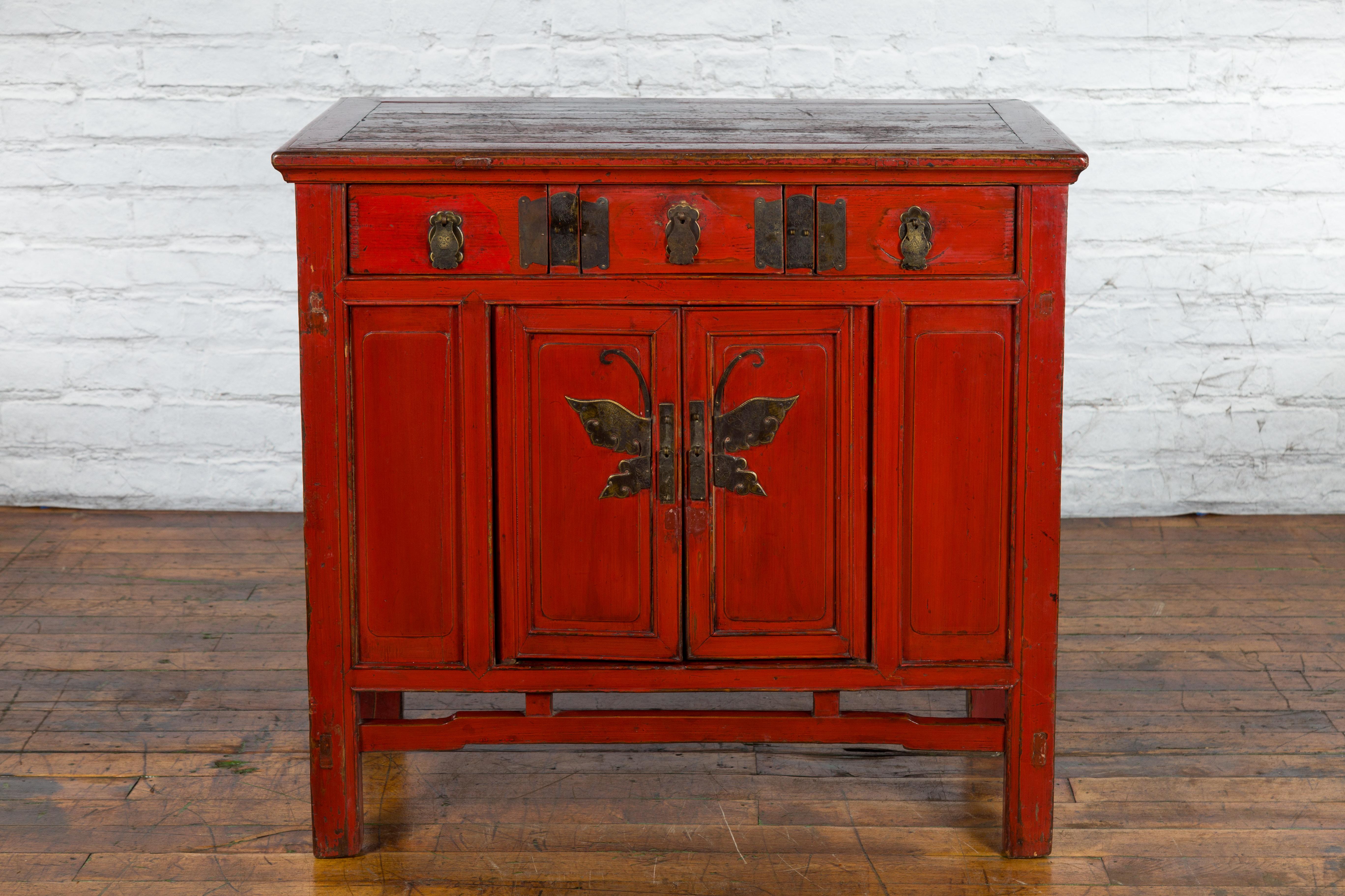 A Chinese Qing Dynasty period cabinet from the 19th century with red lacquer, butterfly bronze hardware, three drawers over two small doors. Created in China during the Qing Dynasty in the 19th century, this cabinet captures our attention with its
