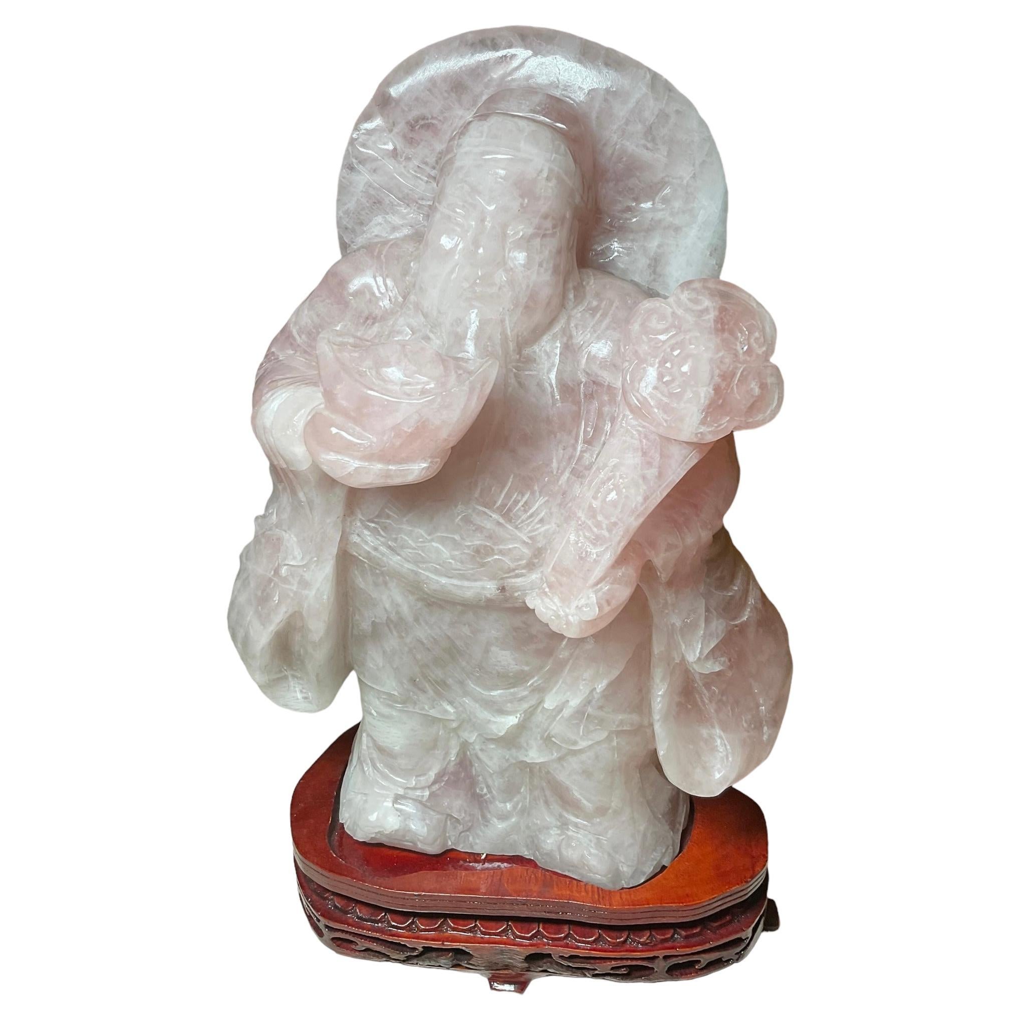 This is a rose quartz heavy sculpture of a Chinese man holding a ruyi scepter. The sculpture is standing over an asymmetrical oval base adorned with some carved scrolls and scalloped motif pattern.