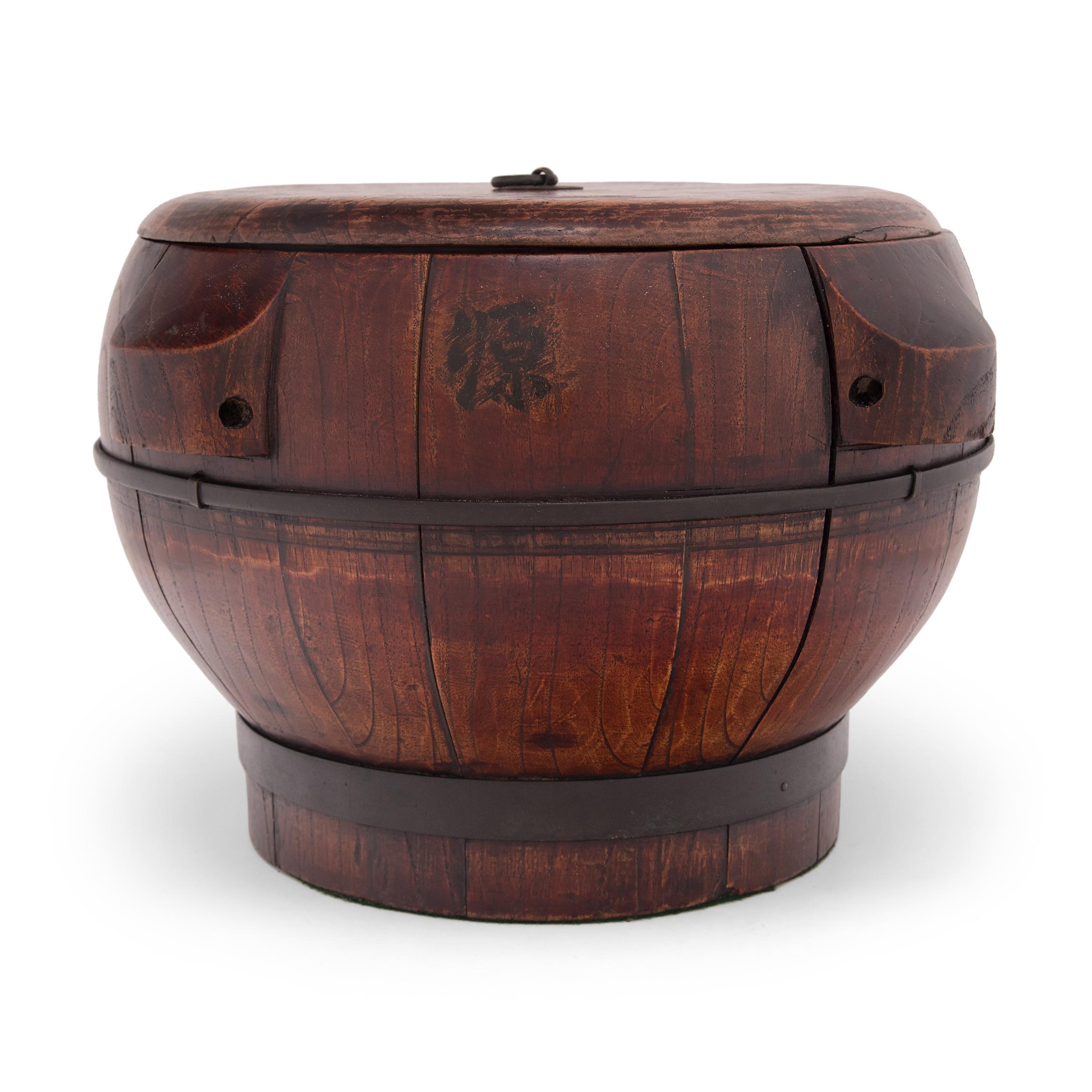This hand-crafted wood container dates to the early 20th century and was used to store rice and other grains. The lidded container has a rounded body with a short footed base, constructed of elmwood staves bound with iron rings. Intended for