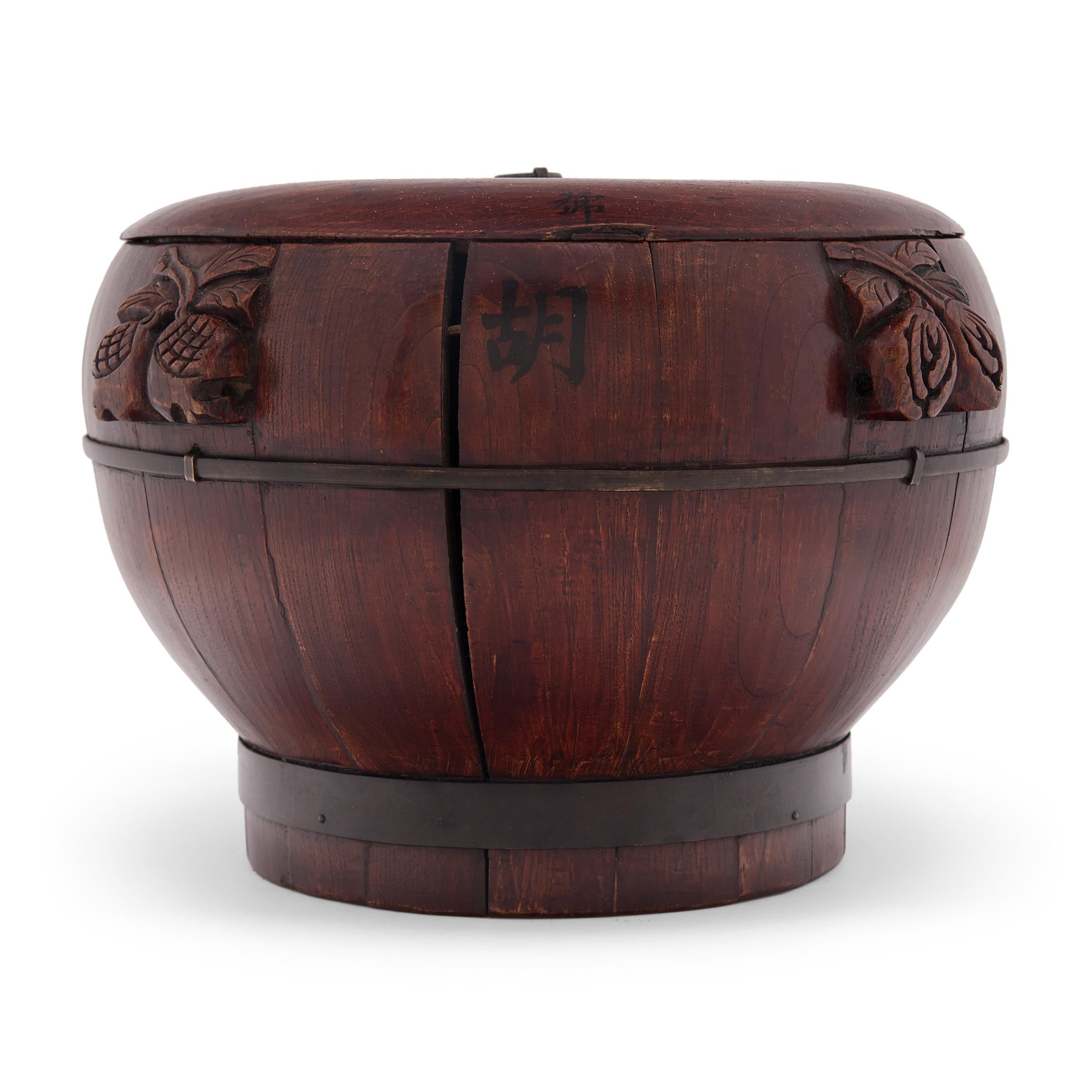 This hand-crafted wood container dates to the early 20th century and was used to store rice and other grains. The lidded container has a rounded body with a short footed base, constructed of elmwood staves bound with iron rings. Darkened to a
