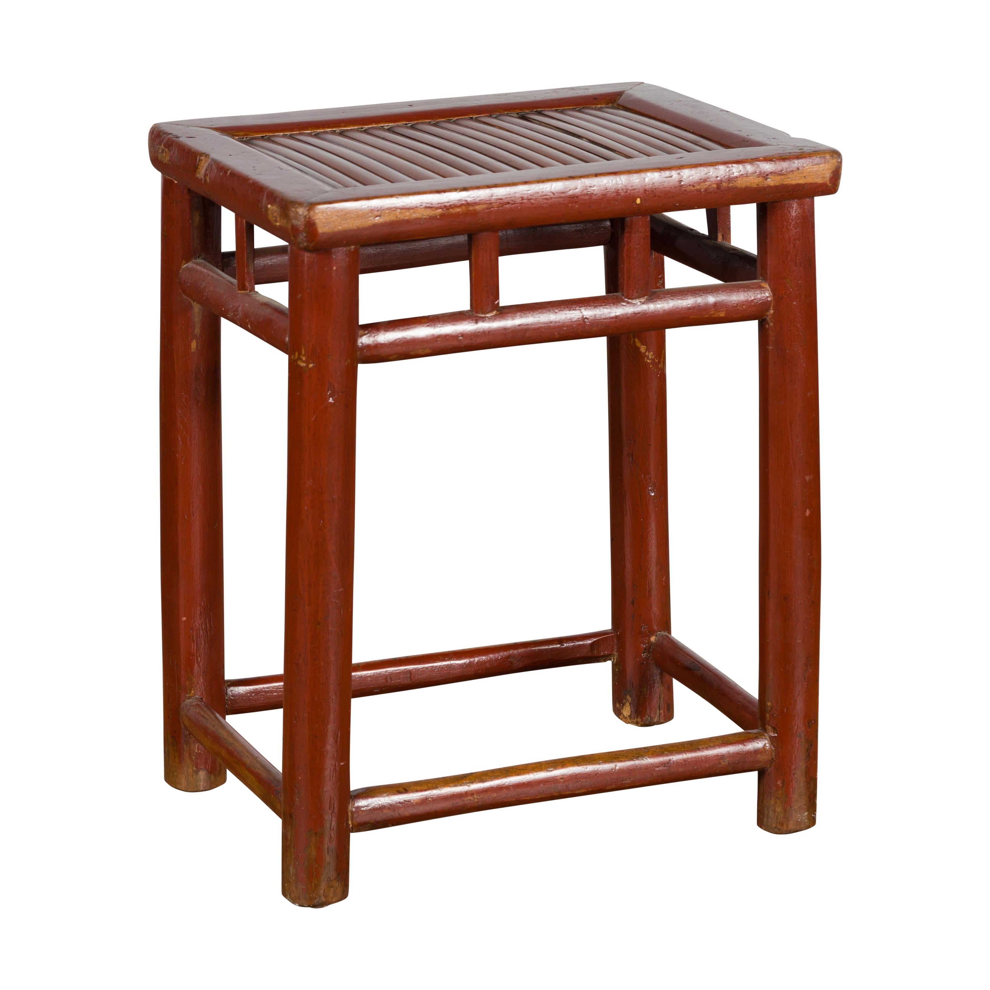 A Chinese reddish brown lacquered stool from the early 20th century, with bamboo seat and pillar strut motifs. Created in China during the early years of the 20th century, this wooden stool features a rectangular top with bamboo seat, resting above