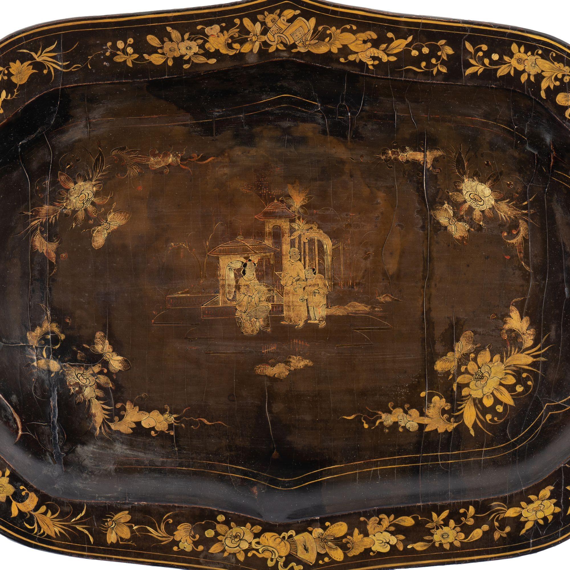 Shaped black lacquer wood tray with gilt decoration. At the center of the design, three figures, one seated and two standing, are positioned in front of two small structures in a garden setting. A repeating and largely symmetrical floral design