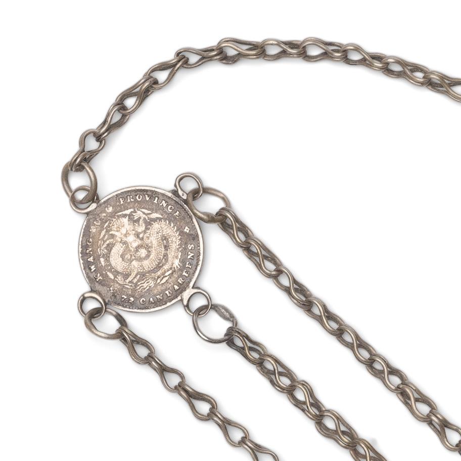 A small Chinese chatelaine tool set strung with a petite white brass tools, including a spoon, a scraper and a pair of tweezers. The small tools hang from short chains linked to a silver Chinese coin originally minted from 1890-1908. Suspended from