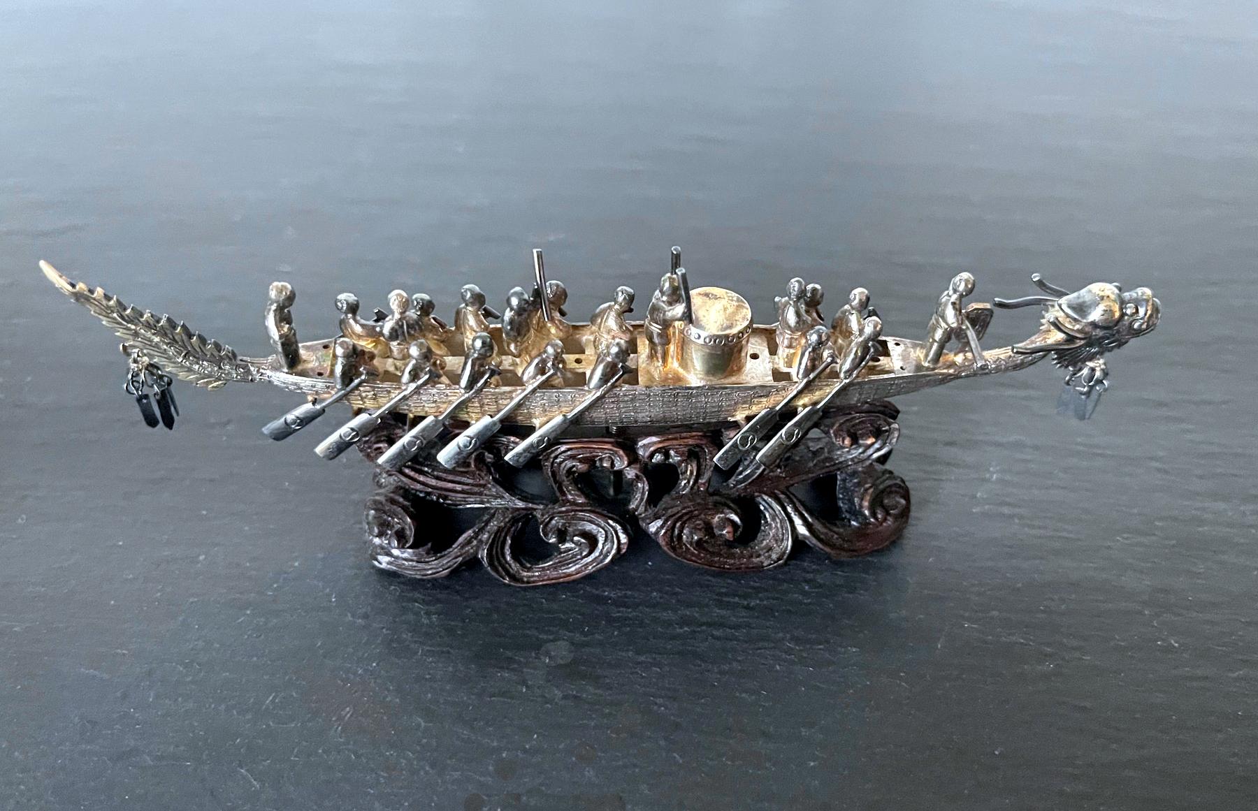 A small Chinese silver cabinet ornamental piece depicting a racing dragon boat with a team of riders displayed on a carved wood stand in the form of waves. The assemblage of riders include a helmsman, a drummer, a cheer leader, a tail man and a team