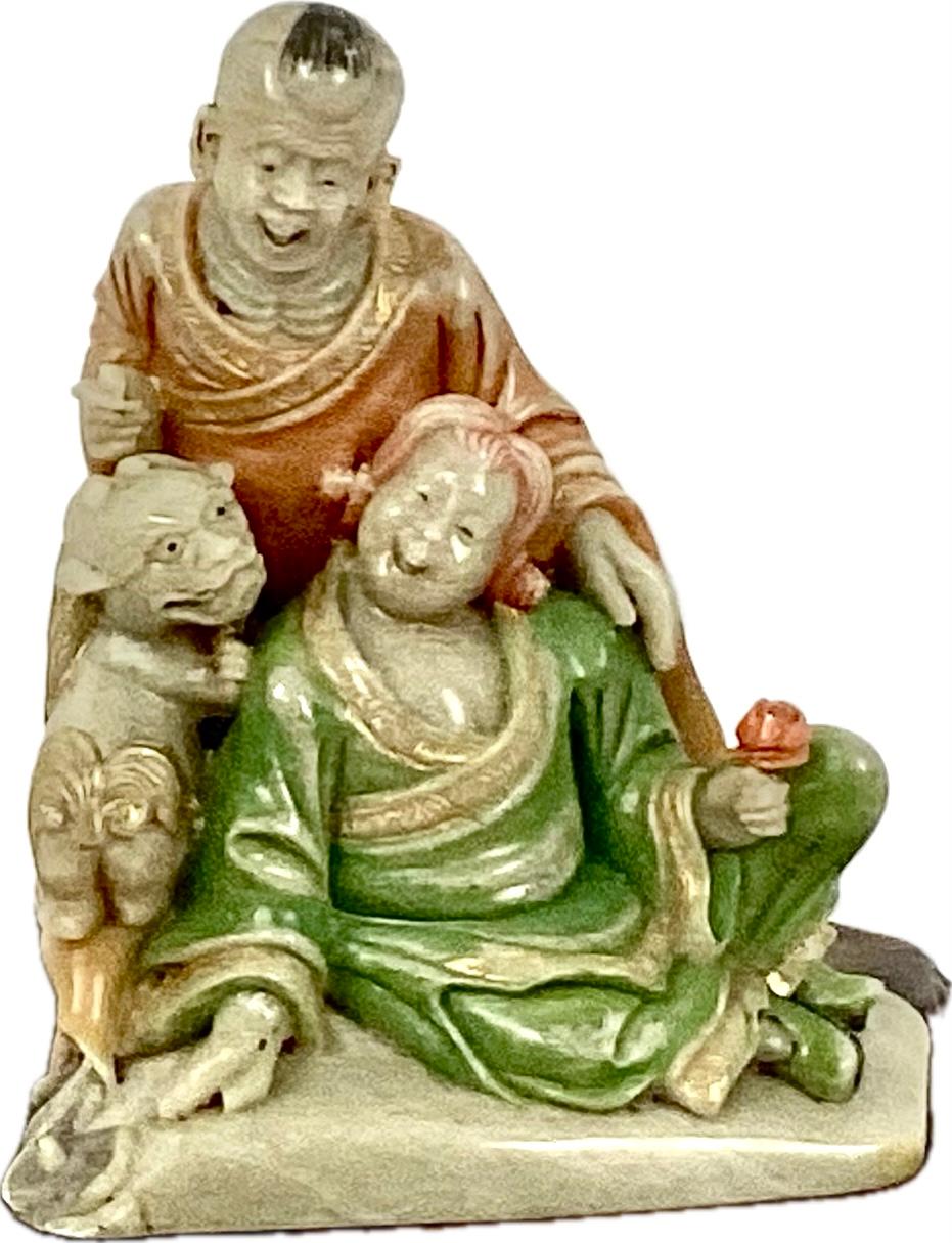 Qing Dynasty soapstone carving depicting a man, a woman and a foo dog sitting and laughing. The woman is holding a red rose and both are wearing colorful Chinese dress in red and green. there is also gold trim throughout. Carving is sitting on a one
