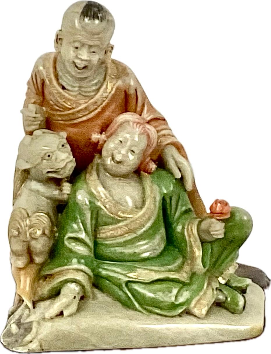 Talla de esteatita de la dinastía Qing que representa a un hombre, una mujer y un perro foo sentados y riendo. La mujer sostiene una rosa roja y ambas visten coloridos trajes chinos en rojo y verde. también hay adornos dorados por todas partes. La
