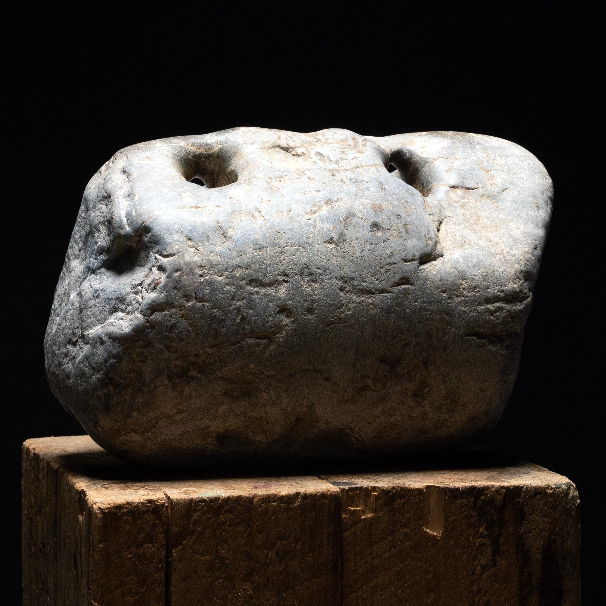 Dépourvu de sculptures figuratives, ce poids en pierre primitif datant du milieu du XIXe siècle présente une beauté organique et abstraite. La pierre a une forme oblongue, en forme de serrure, avec une asymétrie naturelle et deux trous percés sur le