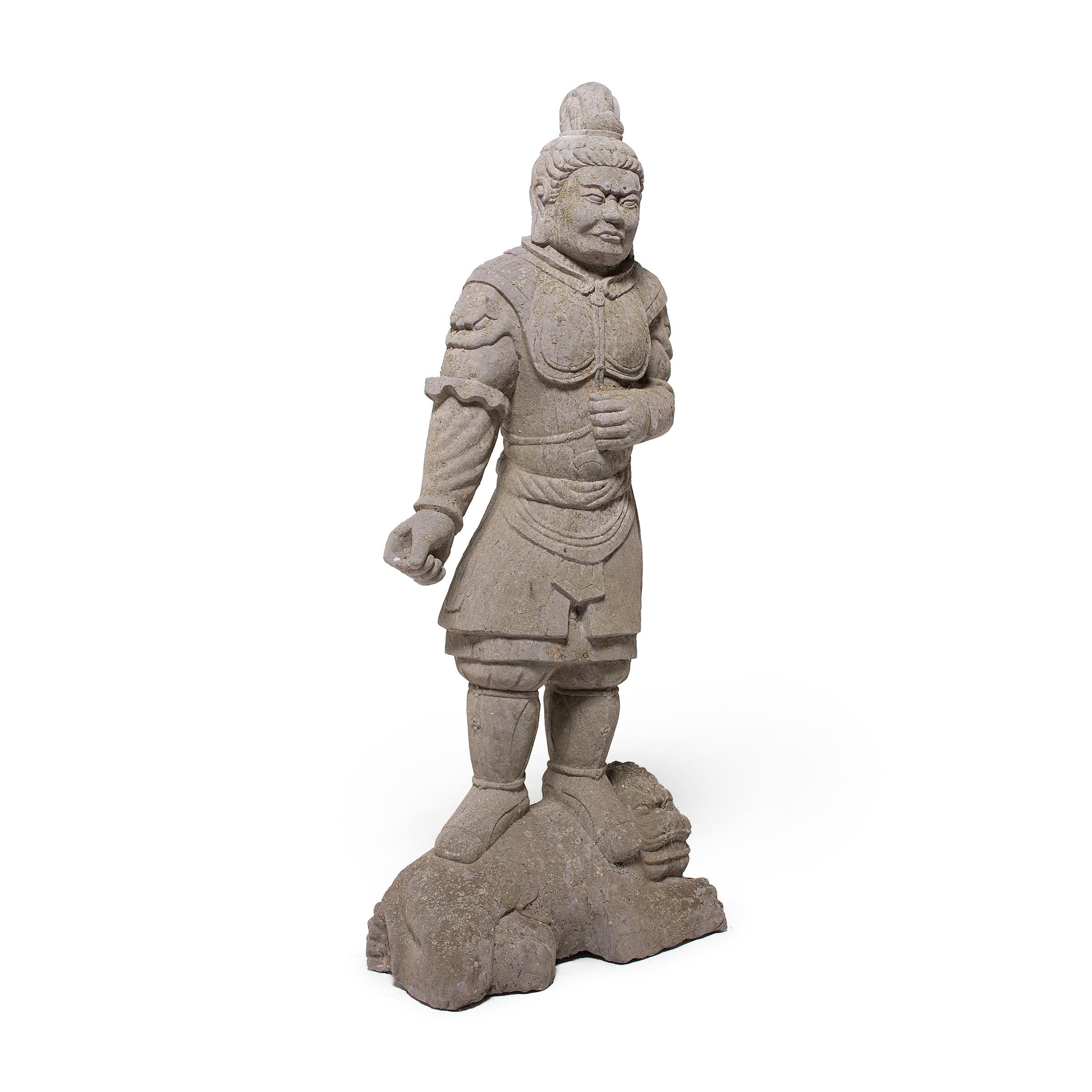This hand-carved stone statue depicts a fearsome Chinese warrior or general, clad in armor with a chest plate and fu dog sleeves. He stands atop a reclining fu dog, or shizi, a posture that symbolizes dominion over the universe. The fu dog is simply