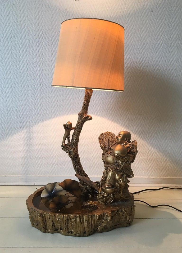 Absolutely stunning and rare table fountain/Lamp. The lamp shows a small boy peeing from a tree and a laughing Buddha sitting at the pond with metal waterlilly's. Base of the lamp was made from some sort of raisin or polystone, while the shade seems