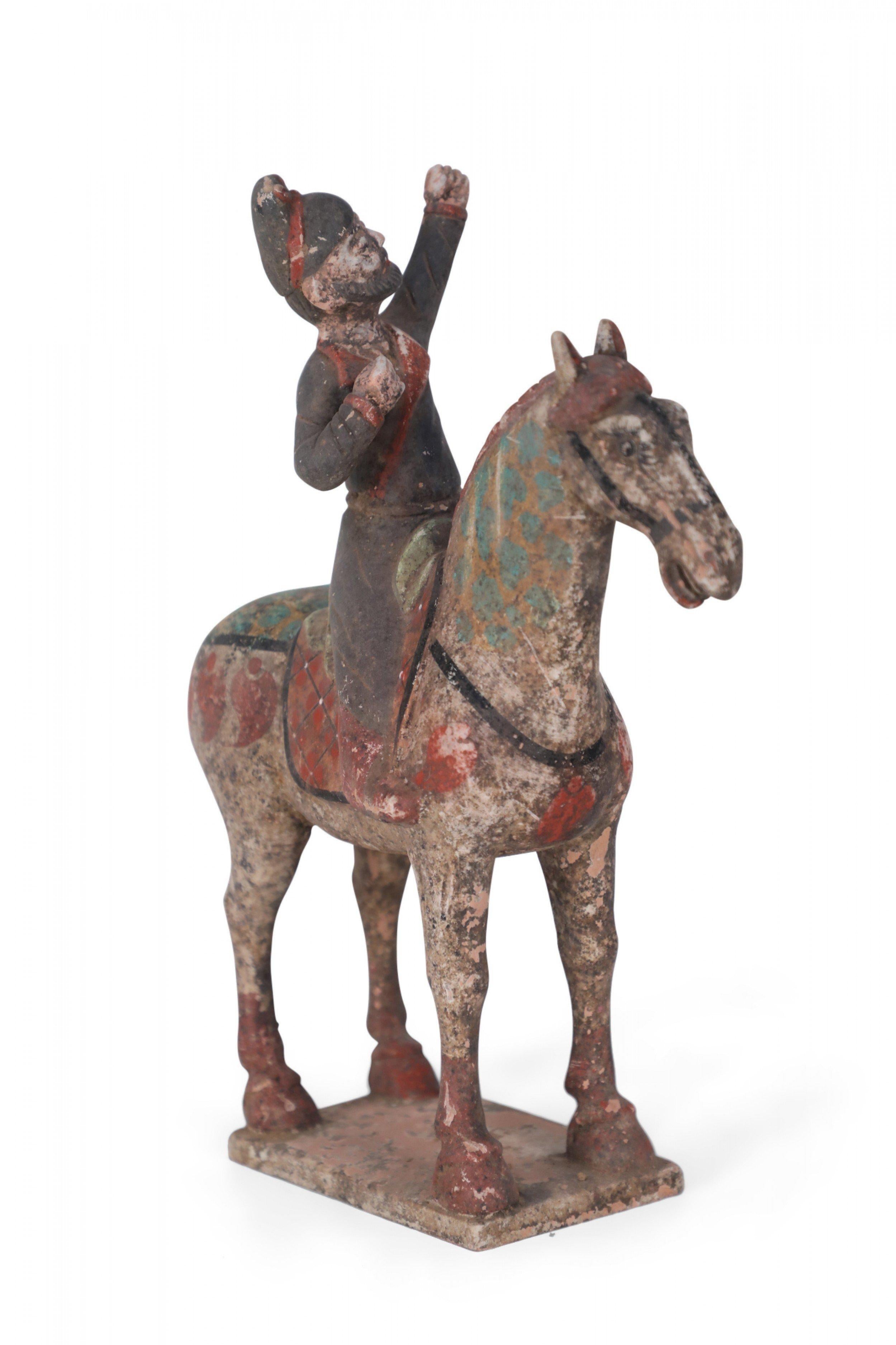 Antique Chinese Tang Dynasty-style terra cotta tomb figure of a man dressed in dark gray robes, with one fist raised in the air, sitting atop a red saddle, on a horse that is painted with blue dots, and standing on a square base.
 