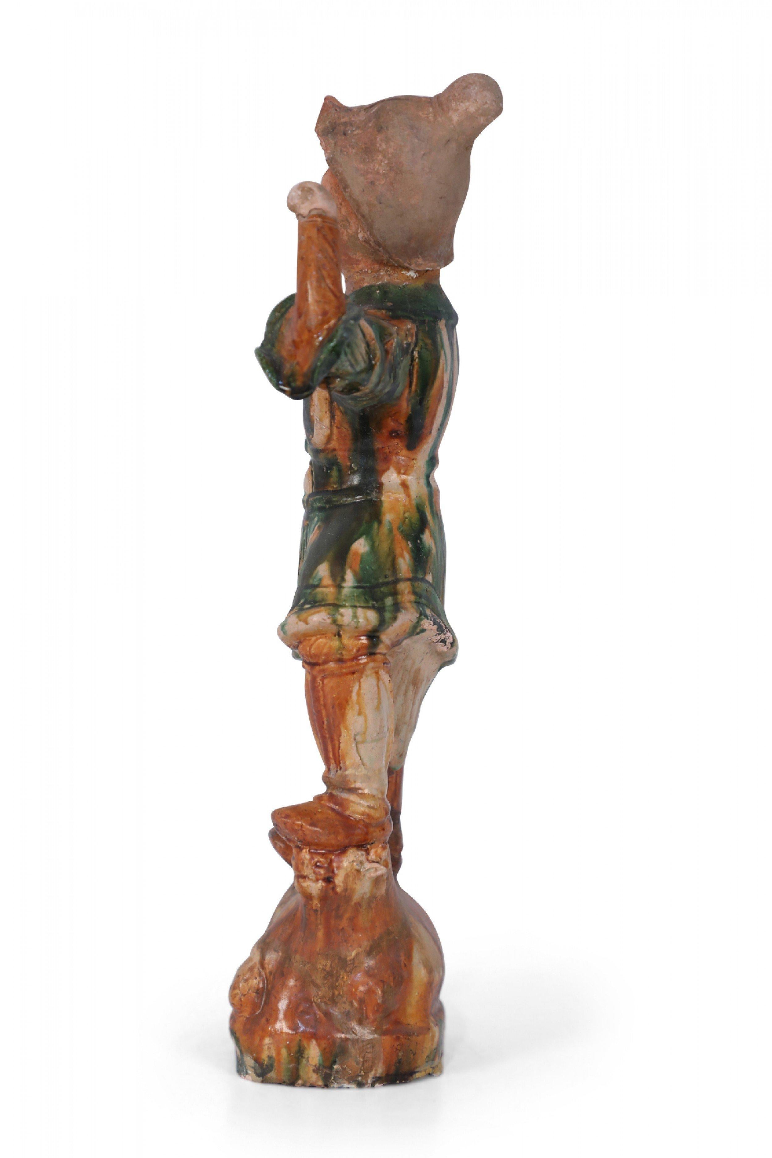 Antigua figura china de terracota, estilo Tang Dynasty, de un guardián sepulcral, en su típica postura de un pie más alto que el otro, apoyado sobre un animal -en este caso, una vaca- y con un brazo levantado, realizada con el tradicional vidriado