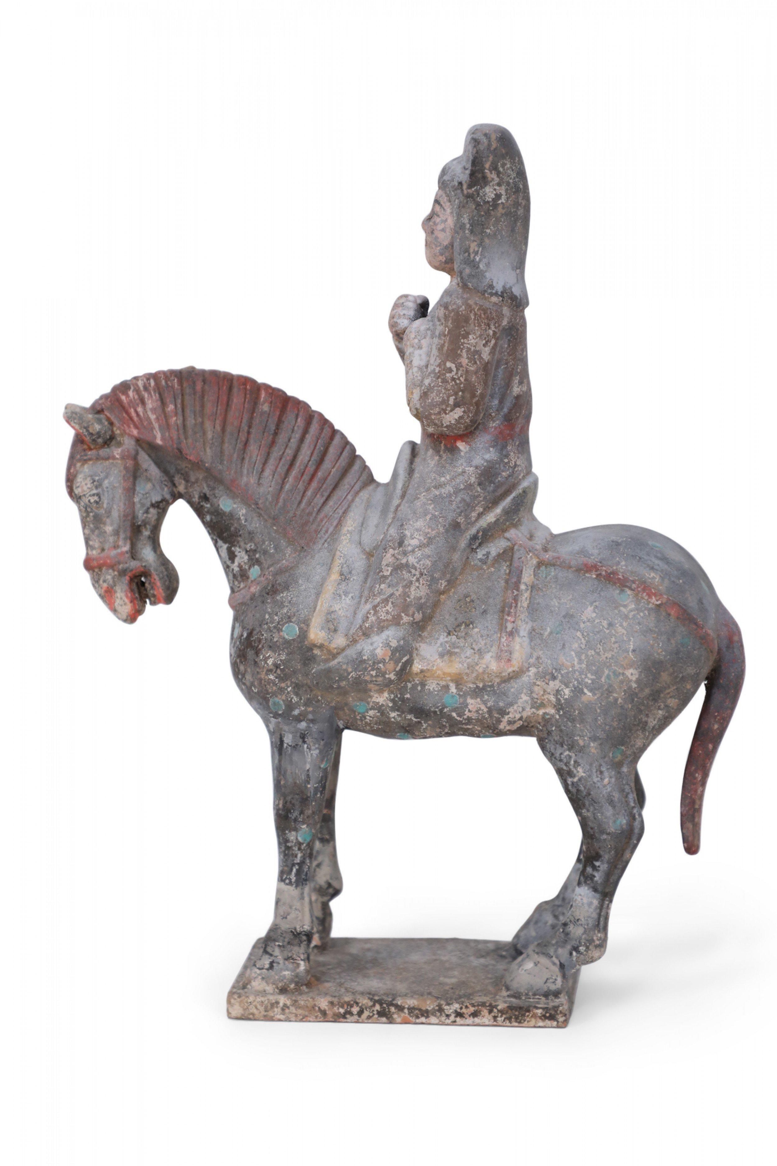 Antique Chinese Tang Dynasty-style terra cotta tomb figure of a rider wearing robes and a red sash sitting on a horse that is painted in blue dots and a red bridle, with its head bowed and standing on a square base.
 