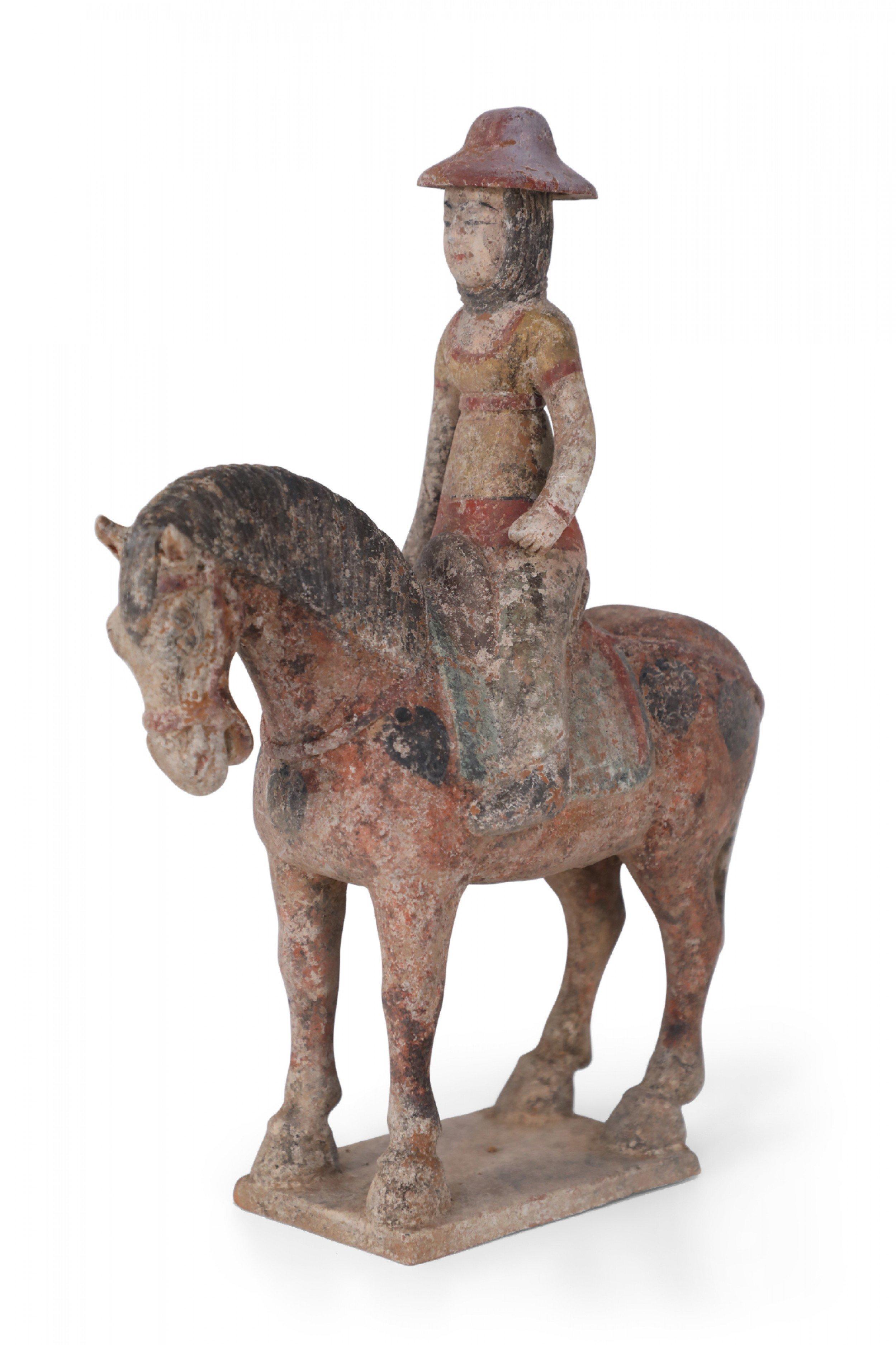 Antique Chinese Tang Dynasty-style terra cotta tomb figure of a girl wearing a red hat riding a brown horse, standing on a textured base.
 