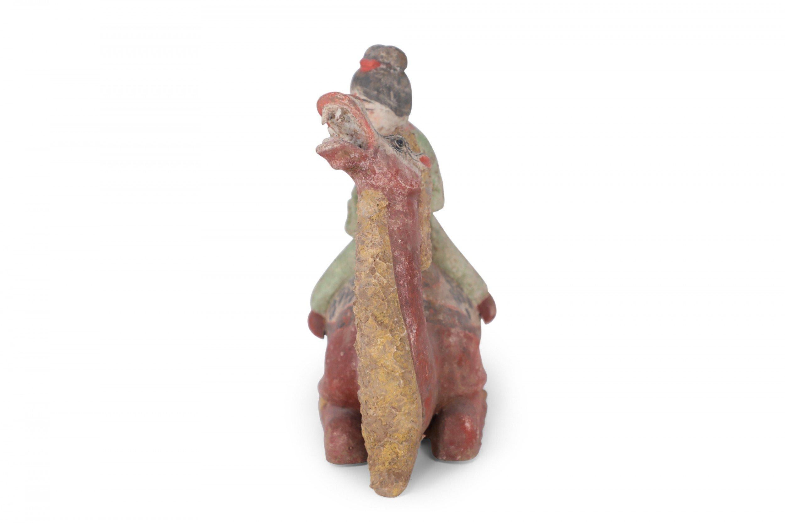 Antique Chinese Tang Dynasty-style terra cotta tomb figure of a woman in a green dress sitting on a kneeling yellow and red Bactrian camel with its head tilted back and mouth open to the sky.
 