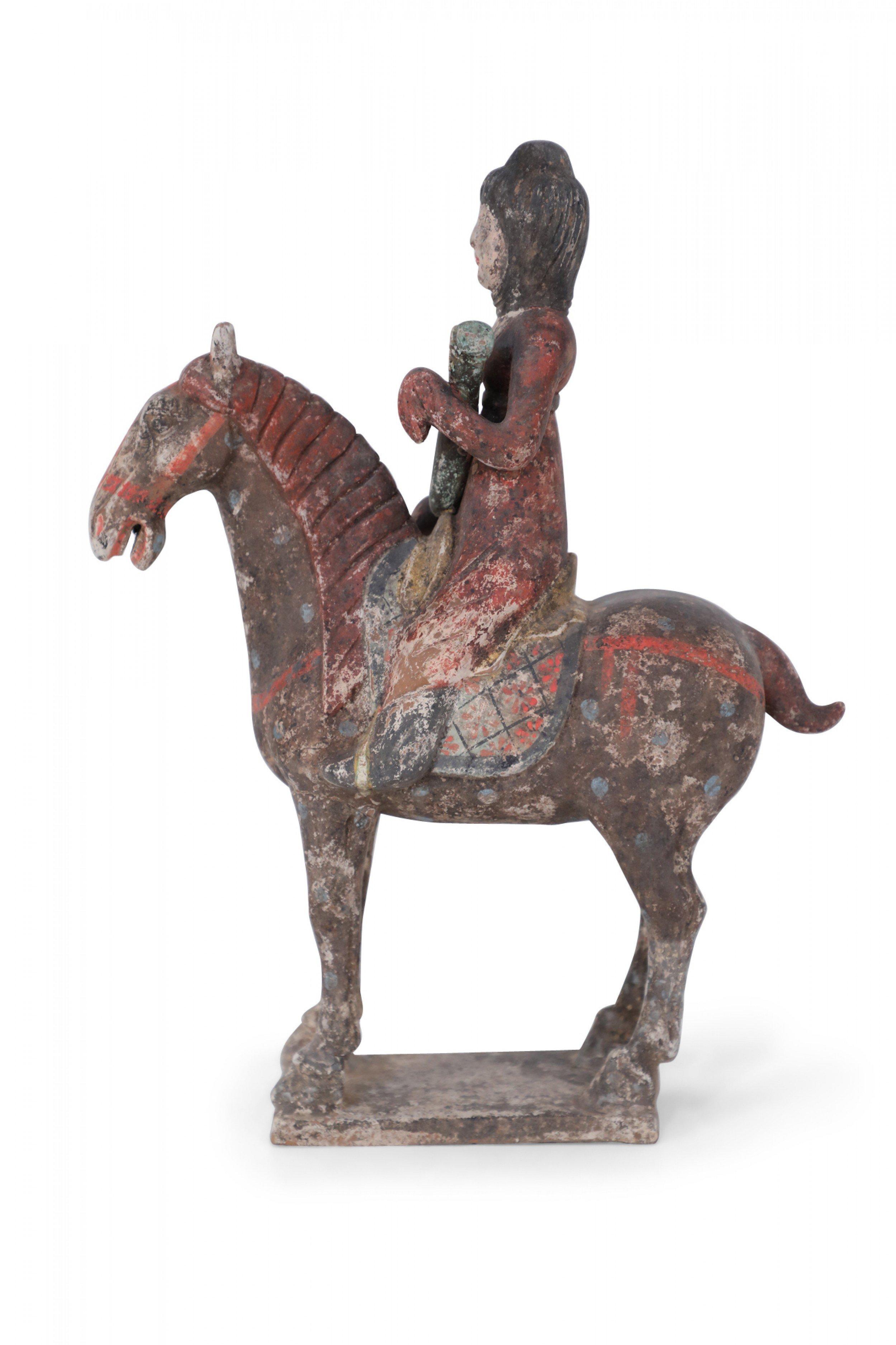 Ancienne figurine funéraire chinoise en terre cuite de style dynastie Tang représentant une jeune fille en vêtement rouge tenant un objet et chevauchant un cheval brun foncé, reposant sur une base texturée.
 
