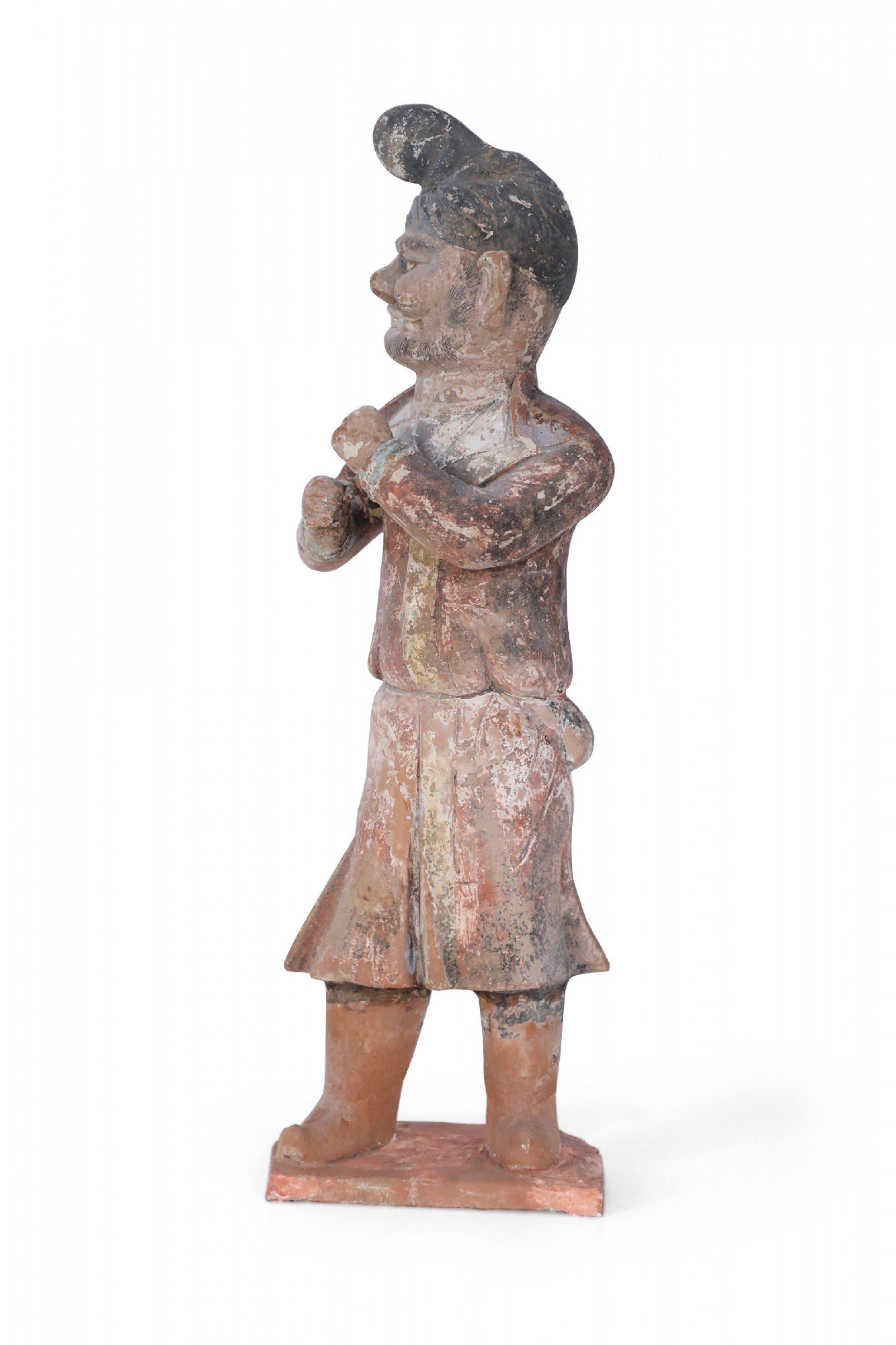 Antique Chinese Tang Dynasty-style terracotta tomb figure of a man with his fists raised, wearing a hat or a head wrap and boots, and standing on a faceted rust-colored base.
 