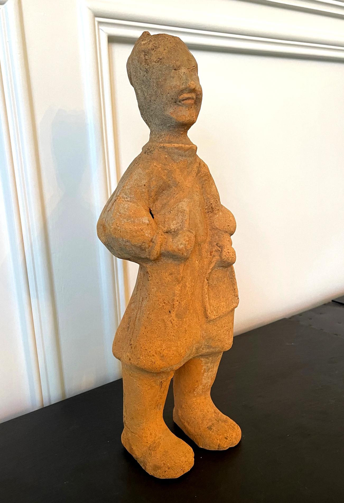 A Chinese terracotta tomb figure (Ni Yong) from East Han Dynasty (25-220 AD), likely from the area of nowadays Sichuan. It depicts a horse groomer with tool and harness in hand. Dressed in a short robe, the figure exhibits a soft smiley facial