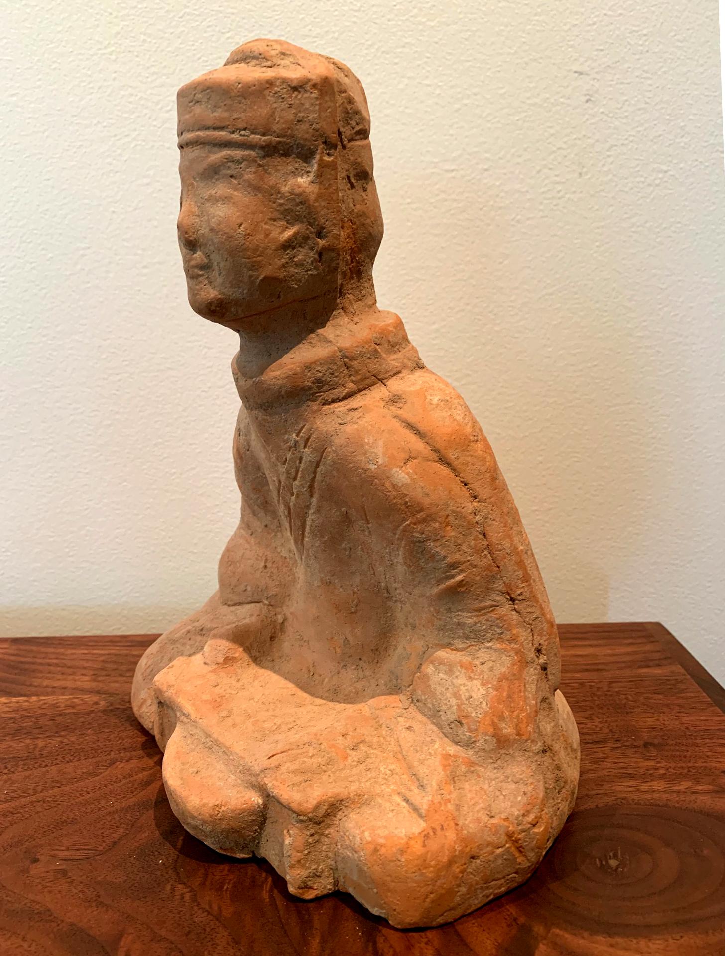 A small Chinese terracotta tomb figure (Ni Yong) from East Han dynasty (25-220 AD), likely from the area of nowadays Sichuan. It depicts a sitting male with courtly attires reading a book. His facial expression shows a faint smile. This kind of