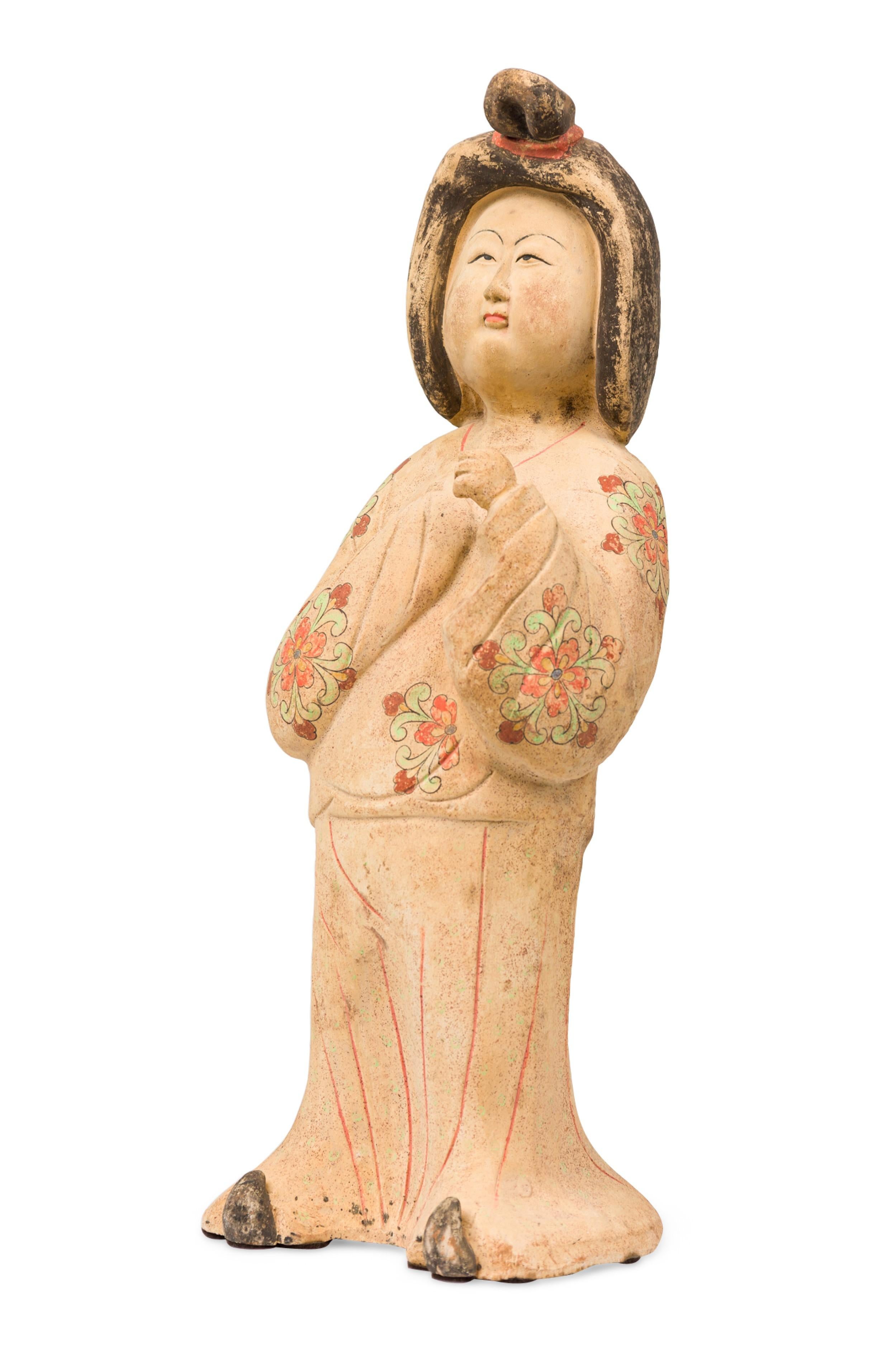 Chinese molded, unglazed beige ceramic figure depicts the stylized Buddhist Goddess posed in a billowy robe with a painted floral pattern, a peaceful smiling facial expression, her left arm raised as if in thought, with upturned slippers peeking out