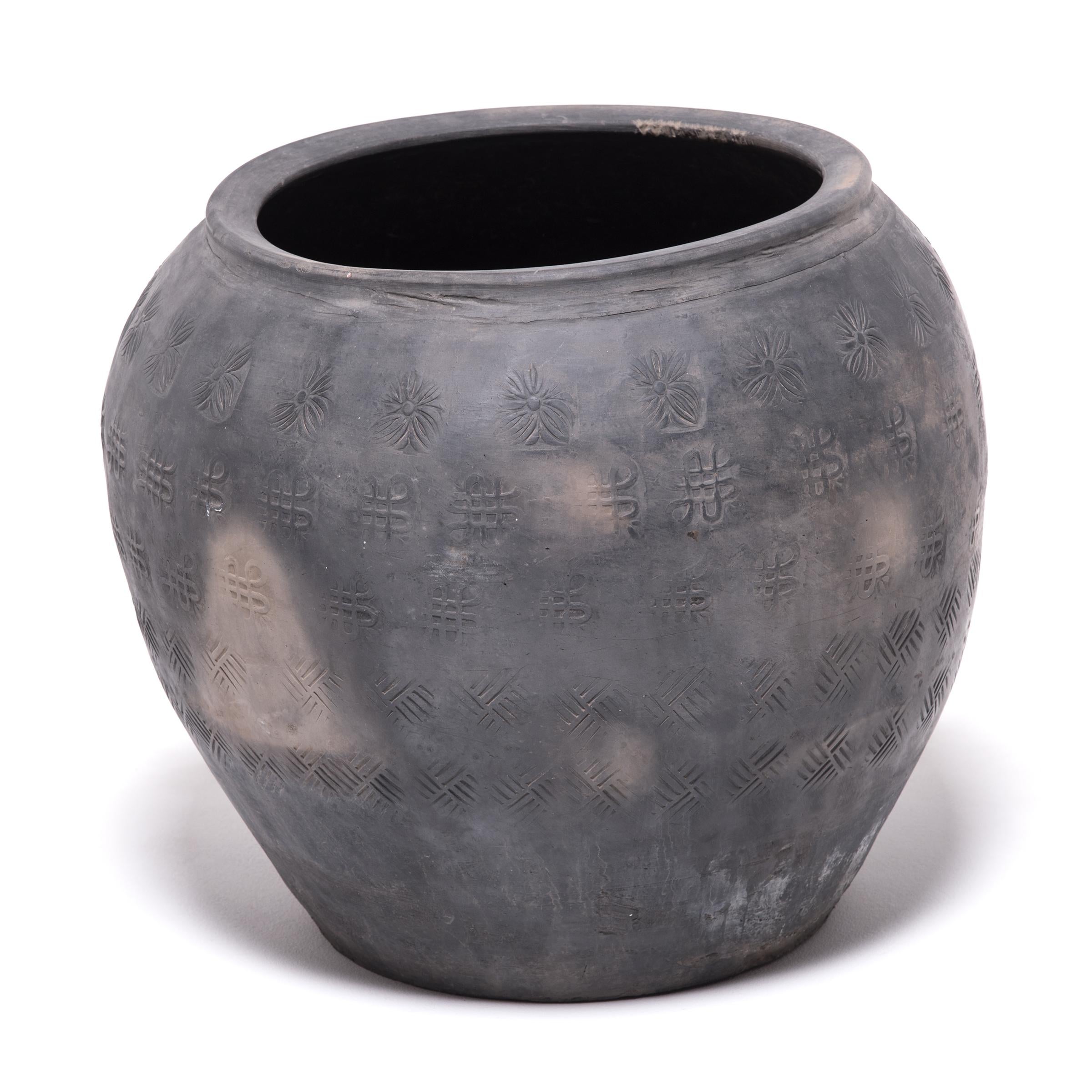 Over a century ago, a gifted potter in northern China crafted this organic clay jar from the rich dark clay of a local river basin. The surface is stamped with auspicious symbols: The Tibetan Buddhist endless knot and the peony, representing Spring